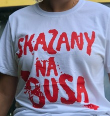 Osoba w białej koszulce z czerwonym napisem 'SKAZANY NA BUSA' i efektem rozprysku farby, widoczna od ramion w górę, na tle pojazdu.