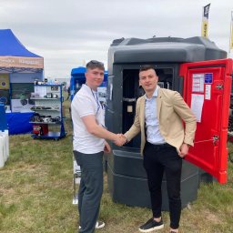 BARTOSZ BUJALSKI - Dwóch mężczyzn w uścisku dłoni na tle przenośnego zbiornika paliwa Kingspan na trawiastym terenie podczas targów, z widocznymi banerami i innymi urządzeniami.