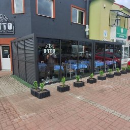 BARTOSZ BUJALSKI - Otto Pizza Napoletana z przeszklonym ogródkiem letnim, stoliki z niebieskimi obrusami w kratę, donice z zielonymi roślinami, czerwony samochód na parkingu.