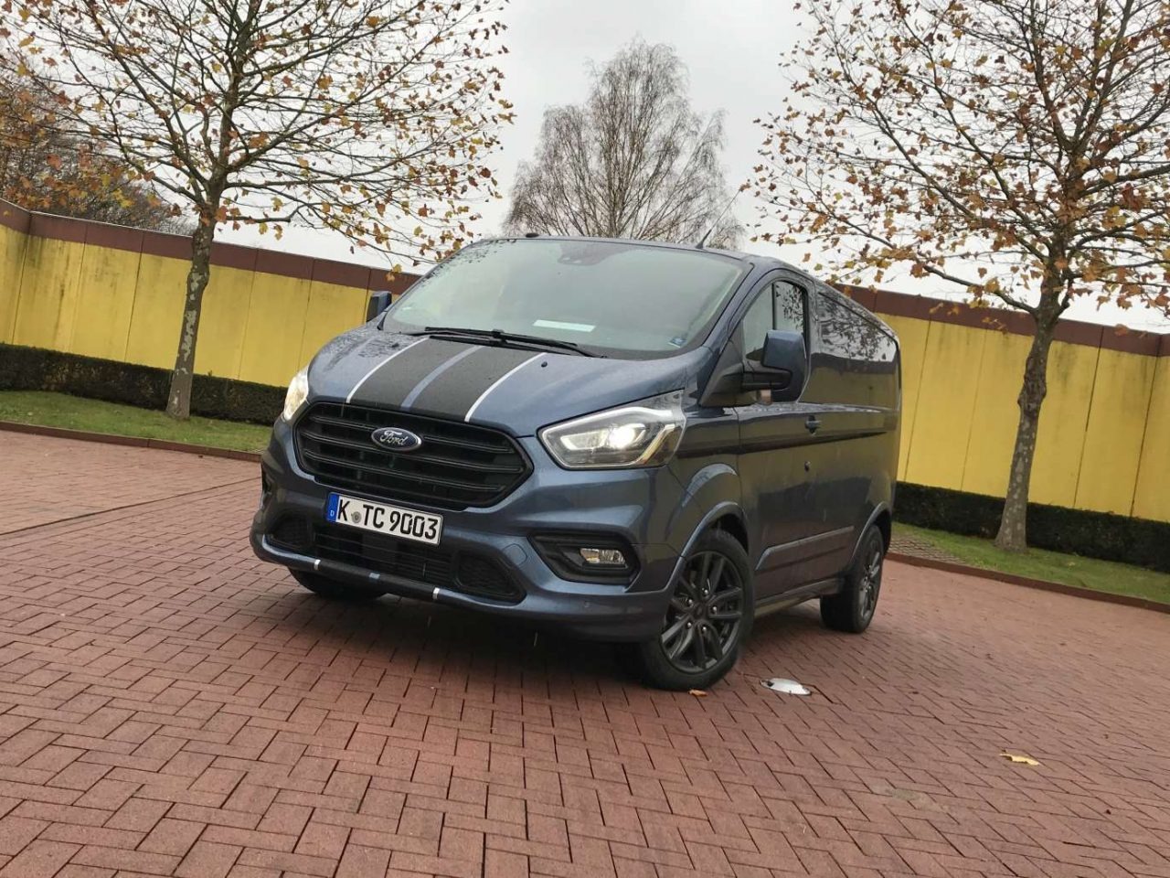 Niebieski Ford Transit Custom z czarnymi pasami i felgami, zaparkowany na brukowanym placu, jesienne drzewa w tle. Tablica rejestracyjna: K TC 9003.