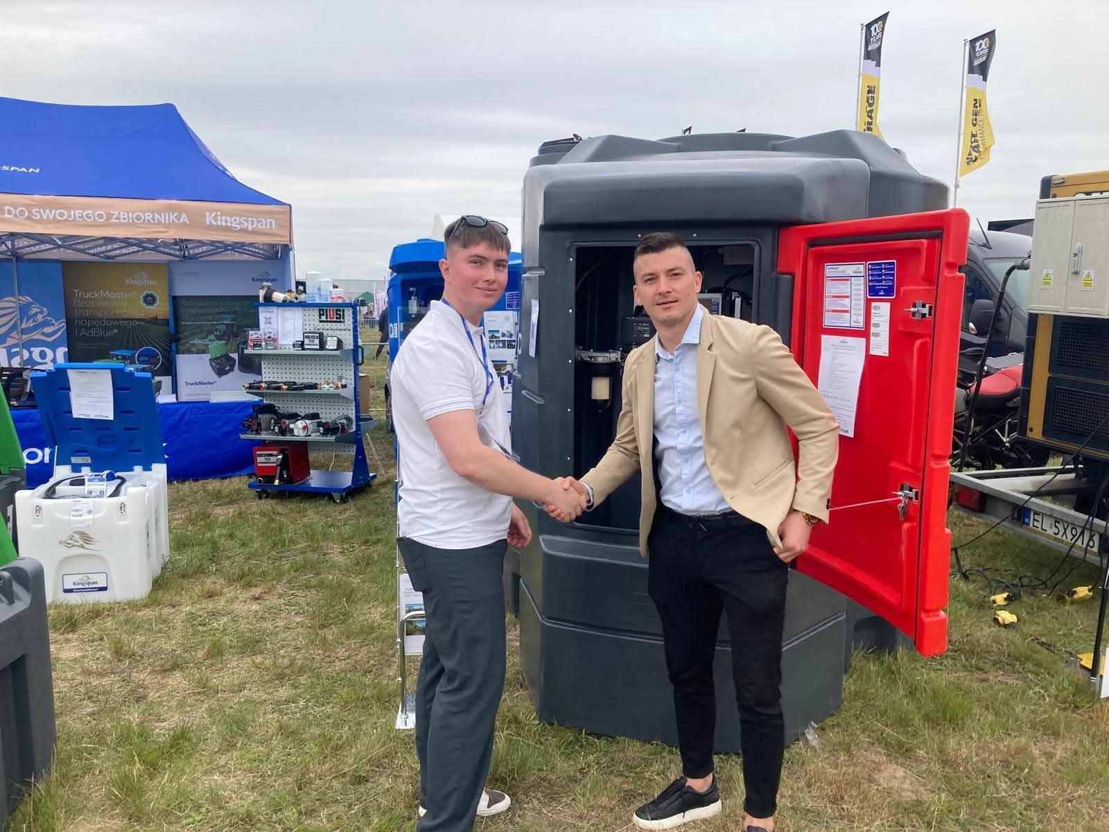 Dwóch mężczyzn w uścisku dłoni na tle przenośnego zbiornika paliwa Kingspan na trawiastym terenie podczas targów, z widocznymi banerami i innymi urządzeniami.