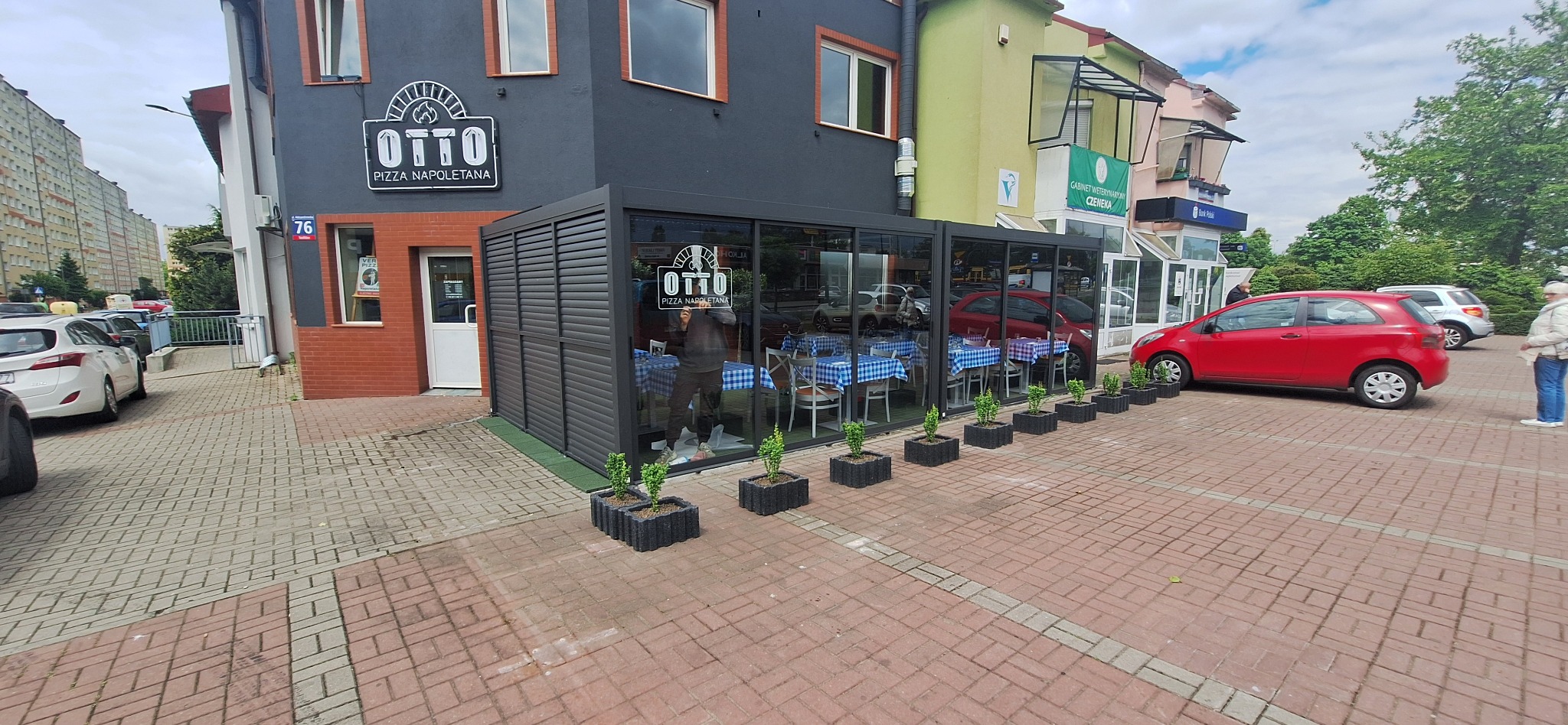 Otto Pizza Napoletana z przeszklonym ogródkiem letnim, stoliki z niebieskimi obrusami w kratę, donice z zielonymi roślinami, czerwony samochód na parkingu.