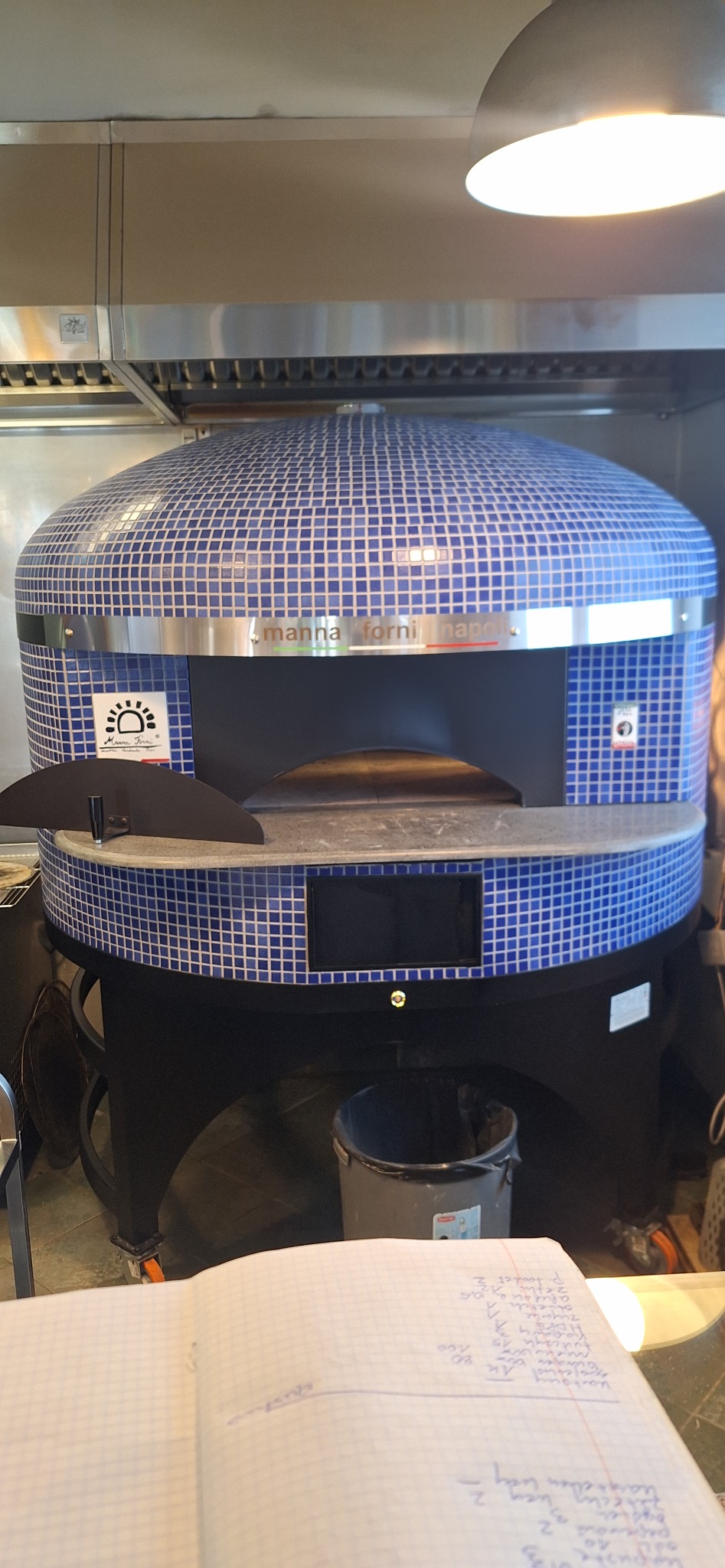 Piec do pizzy Manna Forni Napoli z niebieską mozaiką, stojący w kuchni, z notatnikiem na pierwszym planie. Widoczny fragment wentylacji i lampy.