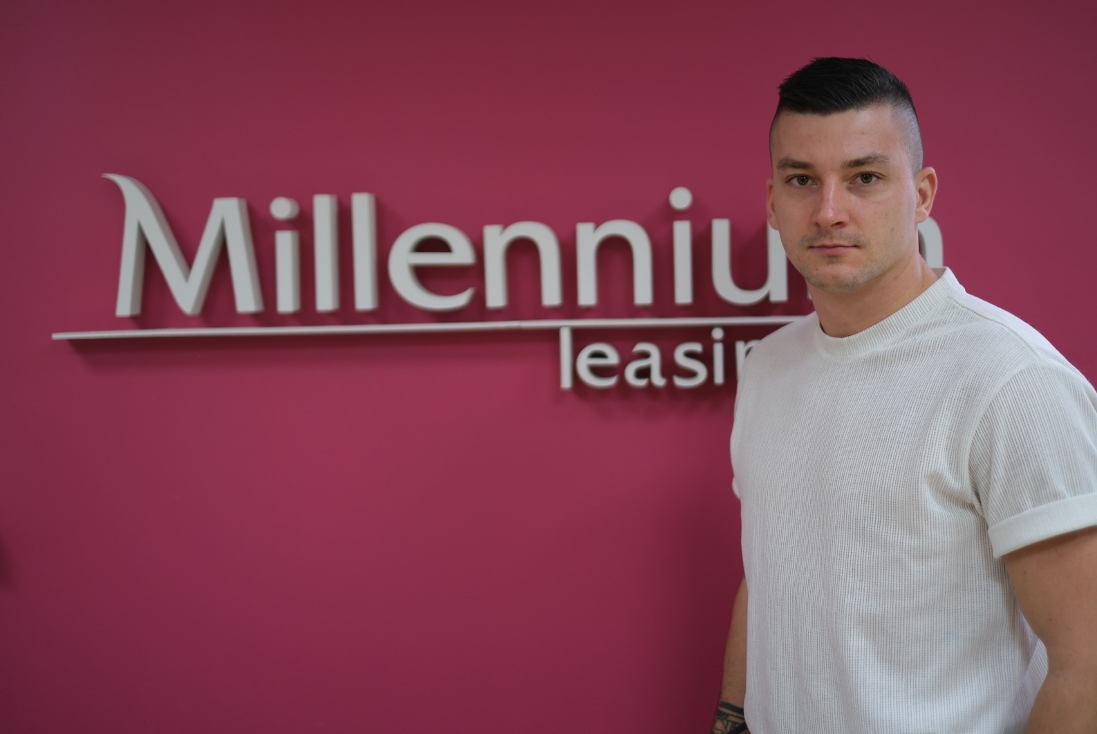 Mężczyzna w białej koszulce na tle różowej ściany z logo 'Millennium leasing'. Ujęcie od pasa w górę, z lekkim przesunięciem w prawo.