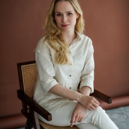 Paulina Dębska photography - Portret kobiety z blond włosami, ubranej w jasną koszulę i spodnie, siedzącej na drewnianym krześle na tle brązowej ściany. Naturalne światło.