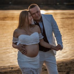 Paulina Dębska photography - Para spodziewająca się dziecka pozuje na tle rzeki i zachodzącego słońca. Kobieta w białej sukience, mężczyzna w białym garniturze. Romantyczna sesja ciążowa.