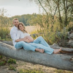 Paulina Dębska photography - Para w jasnych ubraniach siedzi na powalonym drzewie w plenerze. Kobieta w ciąży, mężczyzna obejmuje ją. Romantyczna sesja w naturalnym otoczeniu, w ciepłym świetle.