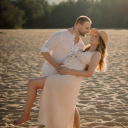Paulina Dębska photography - Para na plaży w ciepłym świetle: mężczyzna trzyma uśmiechniętą, ciężarną kobietę w zwiewnej sukience i kapeluszu. Romantyczna sesja ciążowa na tle piaszczystej plaży i drzew.