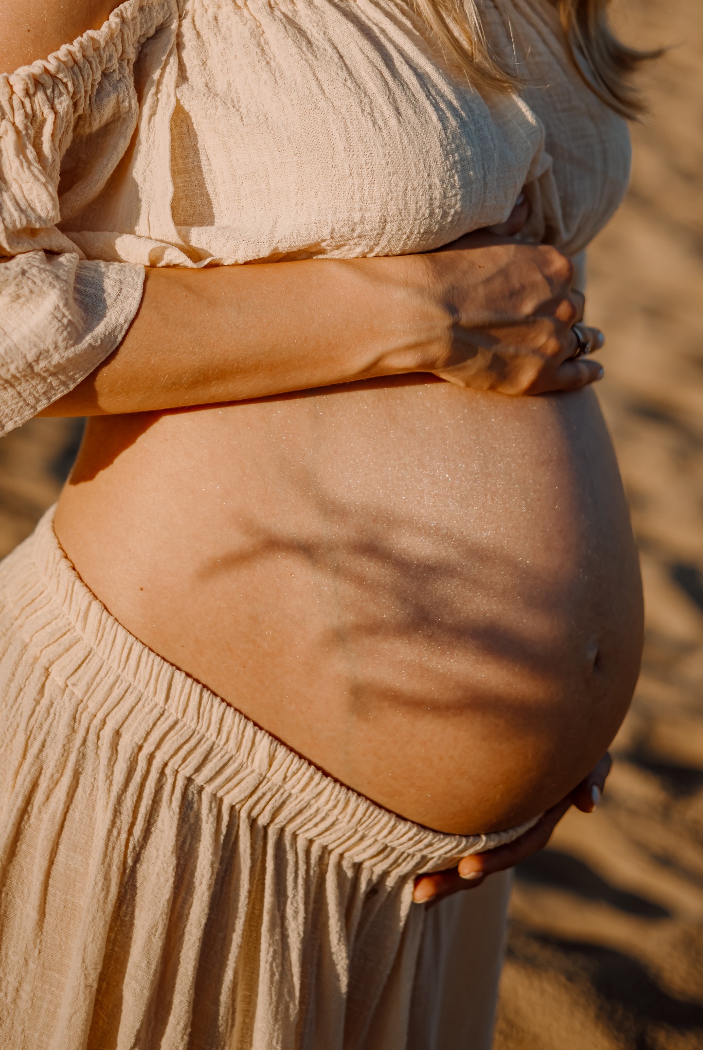 Zbliżenie na ciążowy brzuszek kobiety w beżowej sukience, dłonie obejmujące brzuch, delikatne cienie drzew na skórze, ciepłe światło.