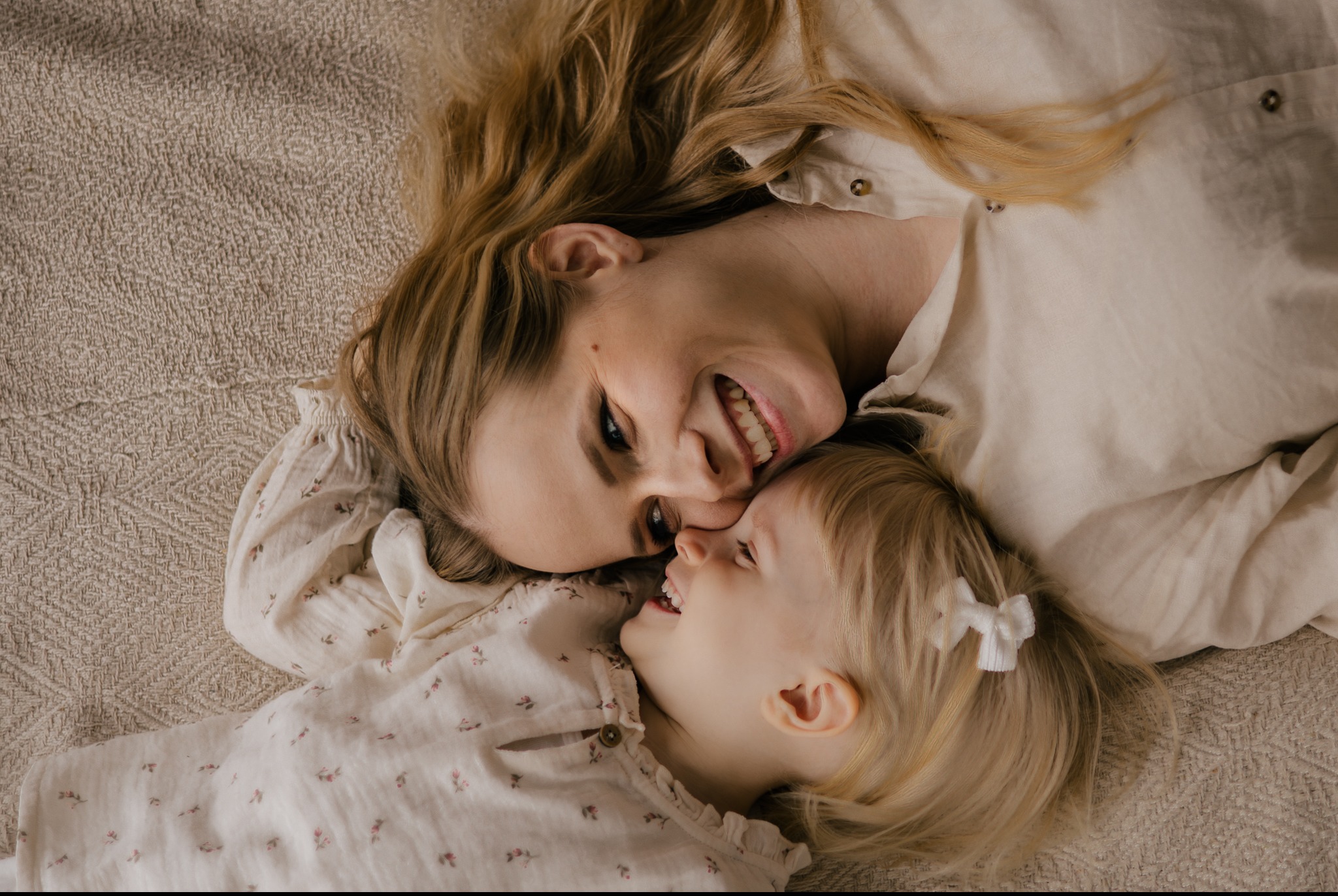 Uśmiechnięta mama i córka leżące blisko na beżowym tle, w jasnych ubraniach z delikatnym wzorem, ujęcie z góry, światło naturalne.