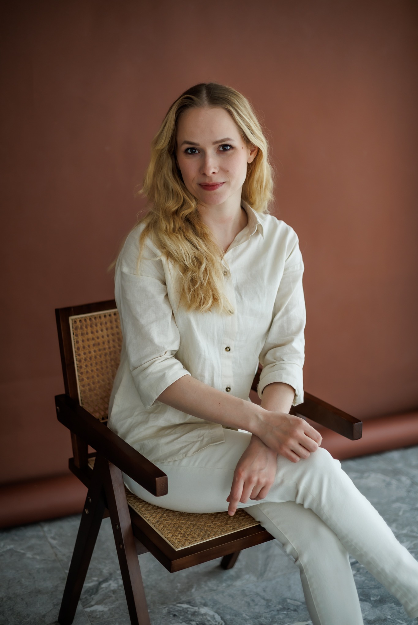 Portret kobiety z blond włosami, ubranej w jasną koszulę i spodnie, siedzącej na drewnianym krześle na tle brązowej ściany. Naturalne światło.