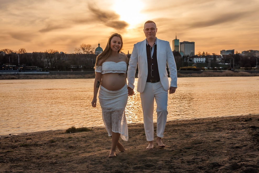 Para na plaży o zachodzie słońca: kobieta w ciąży w białej sukience, mężczyzna w białym garniturze, trzymają się za ręce. W tle rzeka i panorama miasta z wieżowcami.