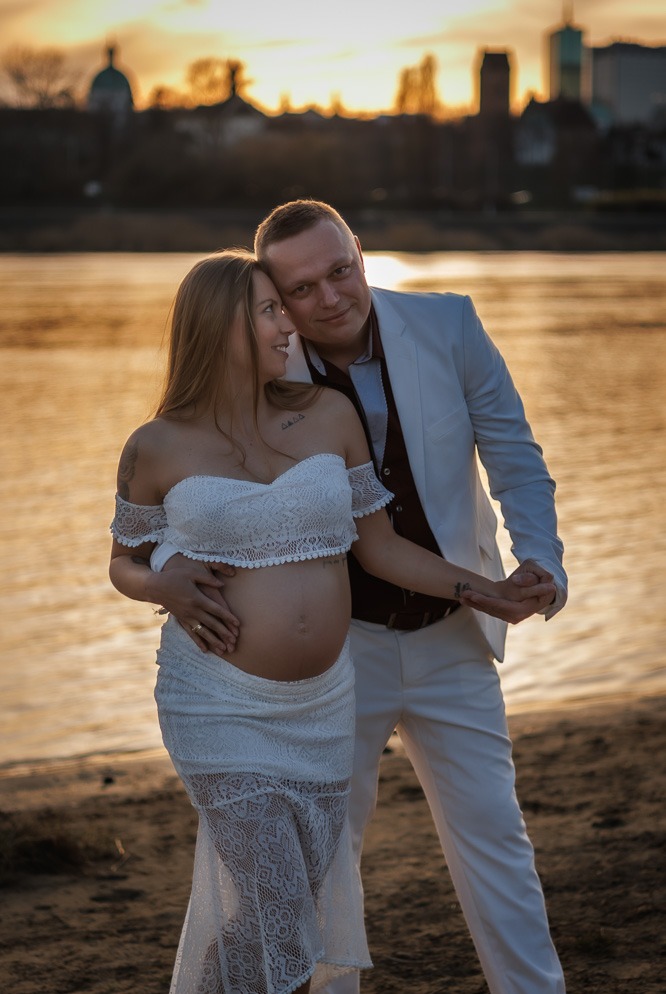 Para spodziewająca się dziecka pozuje na tle rzeki i zachodzącego słońca. Kobieta w białej sukience, mężczyzna w białym garniturze. Romantyczna sesja ciążowa.