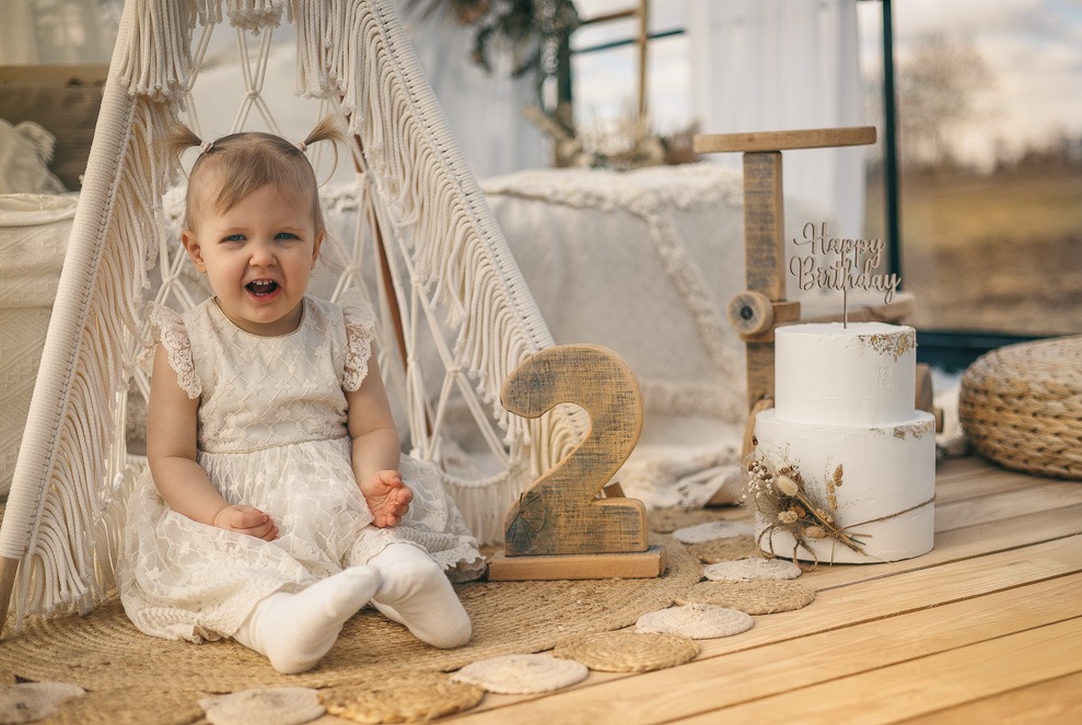 Radosna dwulatka w koronkowej sukience siedzi w tipi obok tortu urodzinowego i drewnianej cyfry '2' na sesji zdjęciowej w stylu boho.