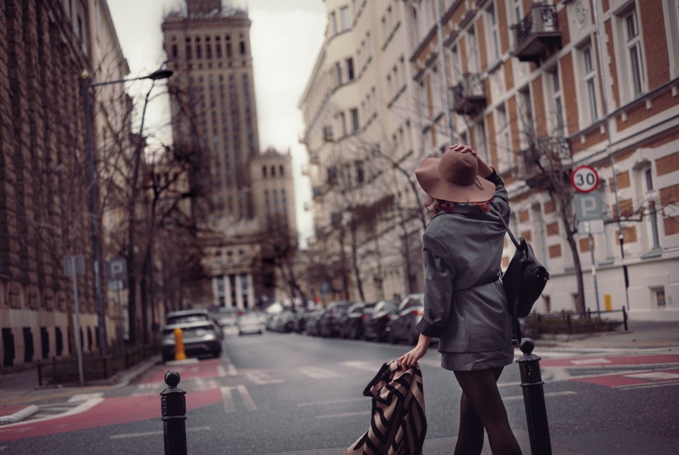 Kobieta w kapeluszu i płaszczu na tle miejskiej ulicy z Pałacem Kultury. Ujęcie od tyłu, styl miejski, jesienna kolorystyka, światło naturalne.