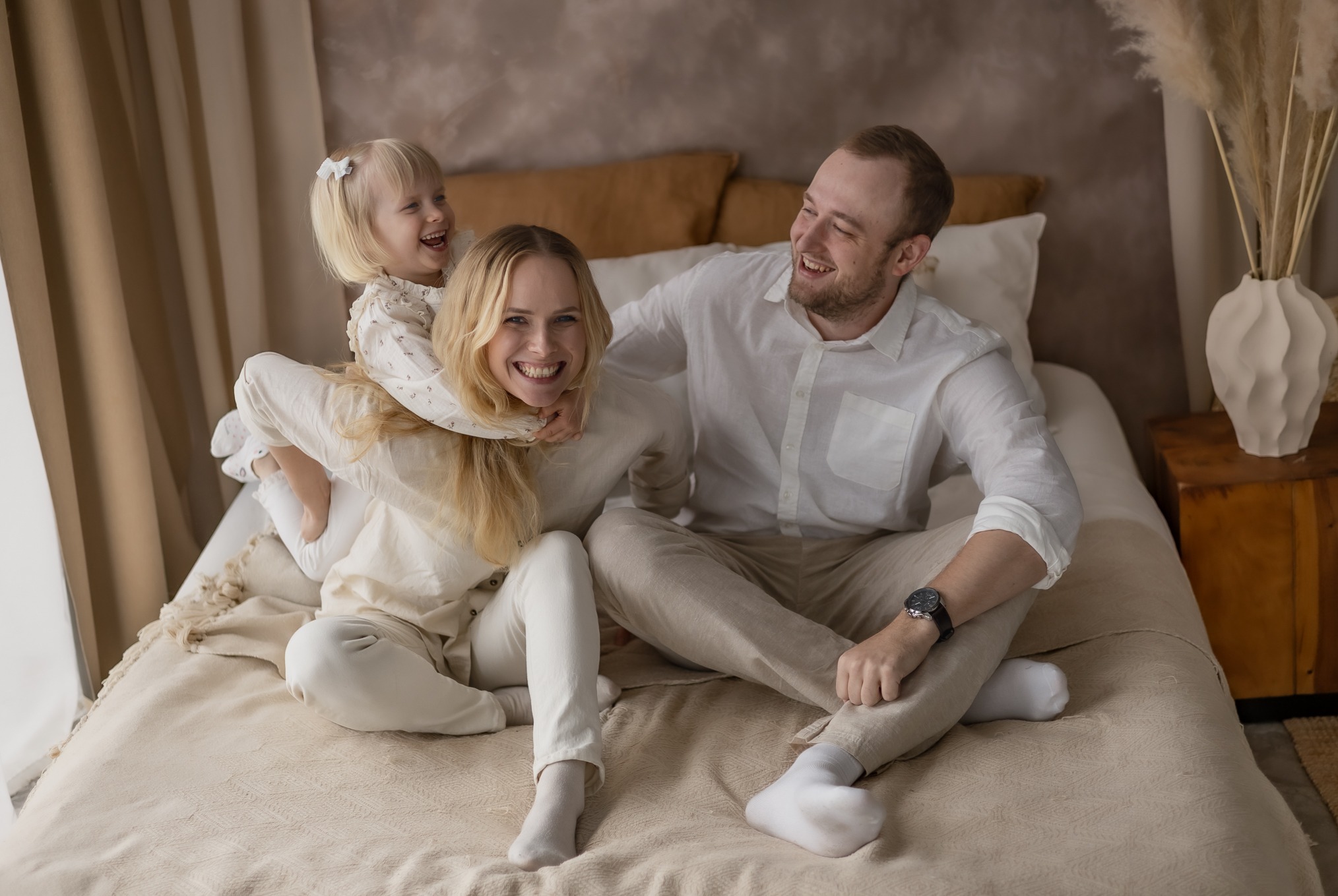 Szczęśliwa rodzina: mama z blond włosami w bieli, tata w koszuli i córeczka na barana, wszyscy uśmiechnięci na łóżku w jasnym wnętrzu. Dekoracyjny wazon i pampasy w tle.