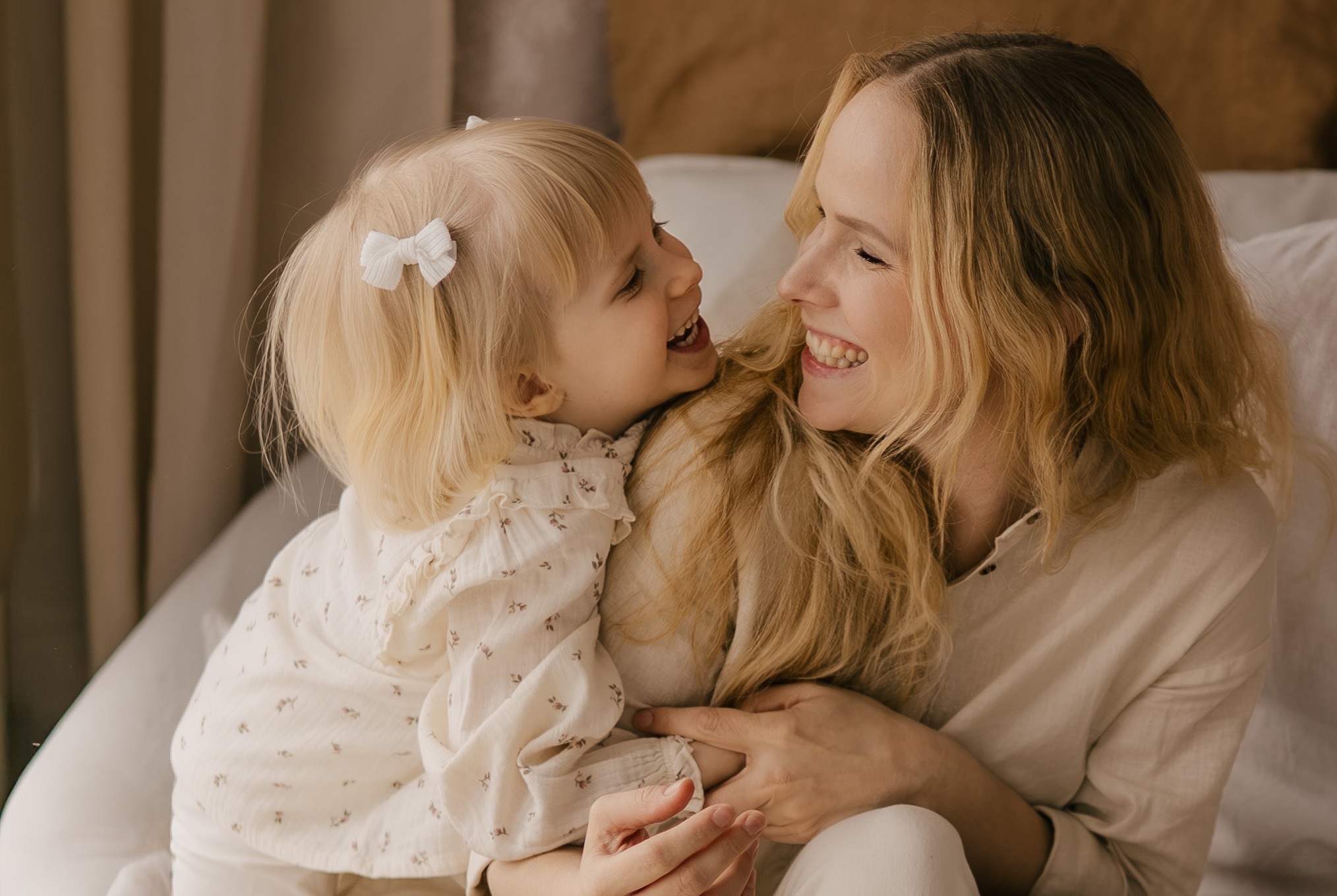 Uśmiechnięta blond mama przytula córeczkę z kokardą we włosach. Jasne, naturalne światło podkreśla ich bliskość i radość. Delikatne wzory na ubraniach dodają uroku.