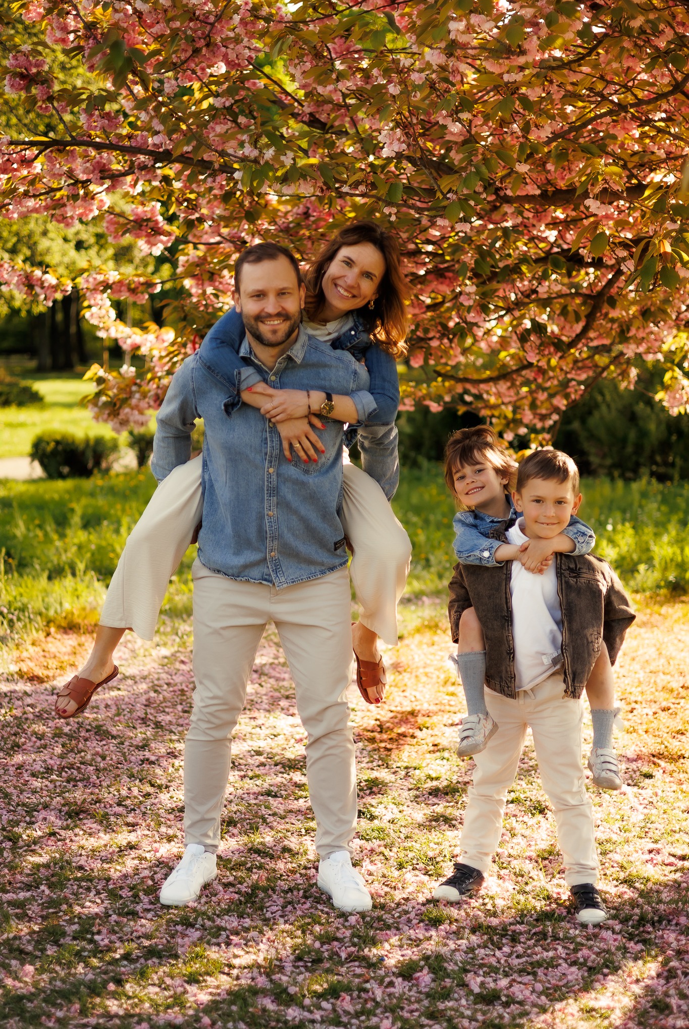 Uśmiechnięta rodzina: mama na barana u taty, syn na plecach drugiego syna, w tle kwitnące drzewo wiśni, park, wiosenna sesja w plenerze, naturalne światło.