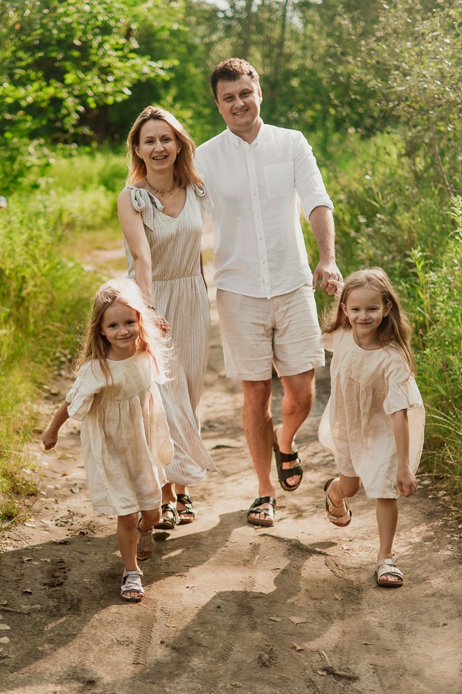 Uśmiechnięta rodzina z dwiema córkami w pasujących, jasnych strojach spaceruje polną drogą w słoneczny dzień. Naturalna sesja rodzinna w plenerze.