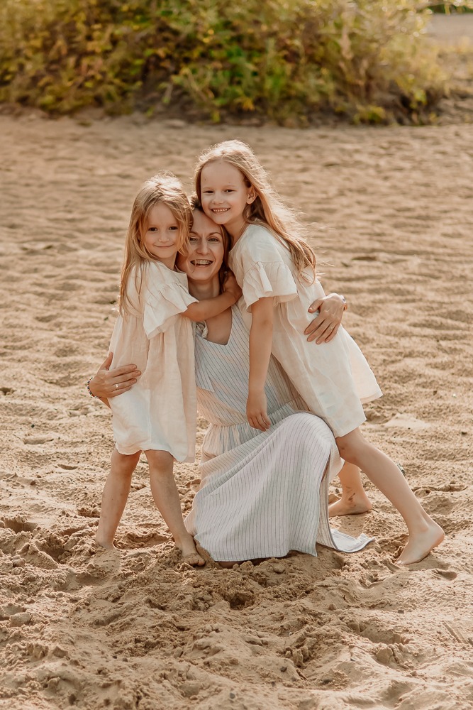 Uśmiechnięta mama w długiej sukience przytulana przez dwie córki na piaszczystej plaży w słoneczny dzień. Dziewczynki w identycznych, jasnych sukienkach.