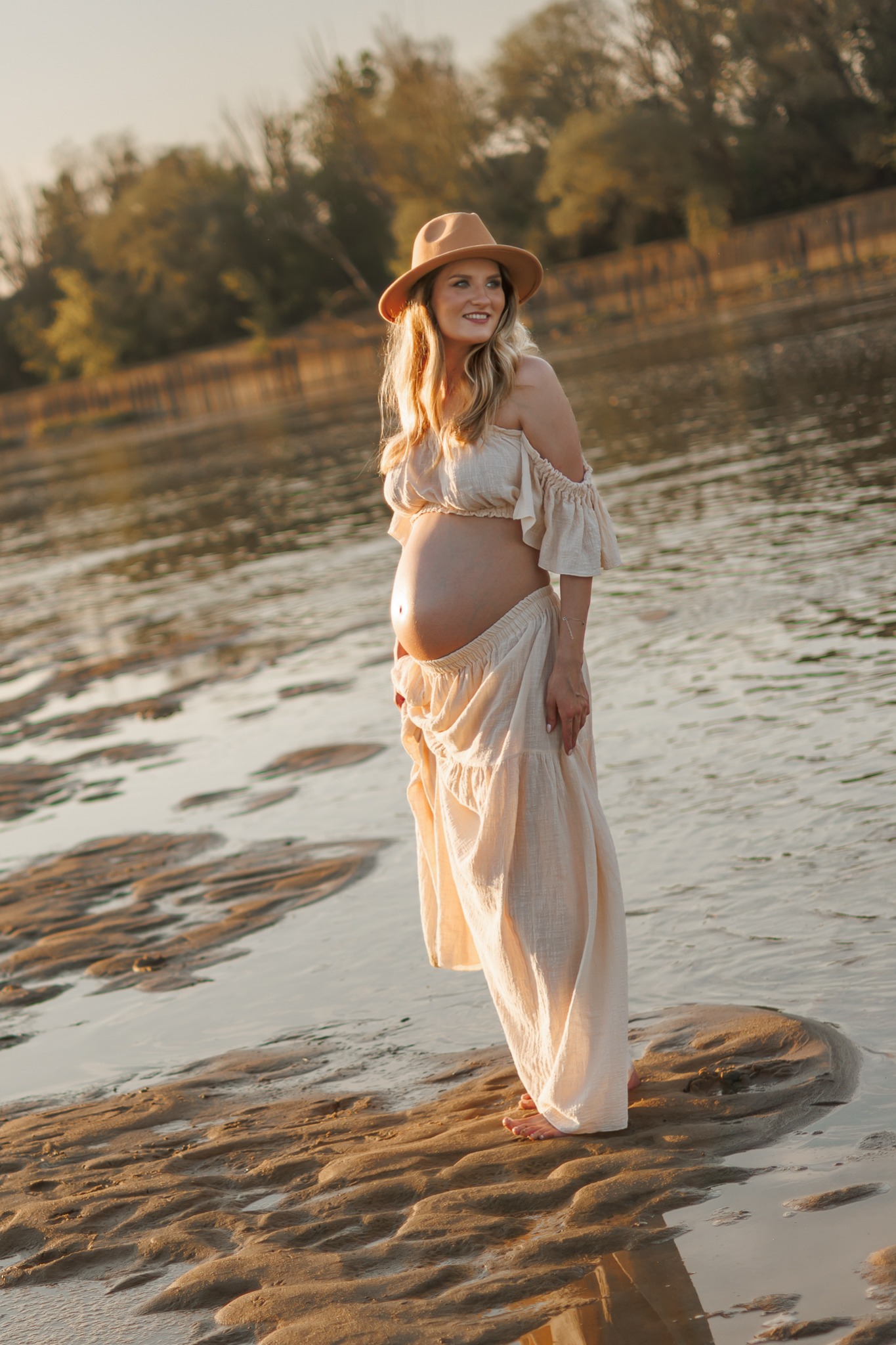 Sesja ciążowa w Warszawie: Kobieta w zwiewnej sukience i kapeluszu stoi na piaszczystej łasze, z wodą w tle, uśmiechając się w bok.