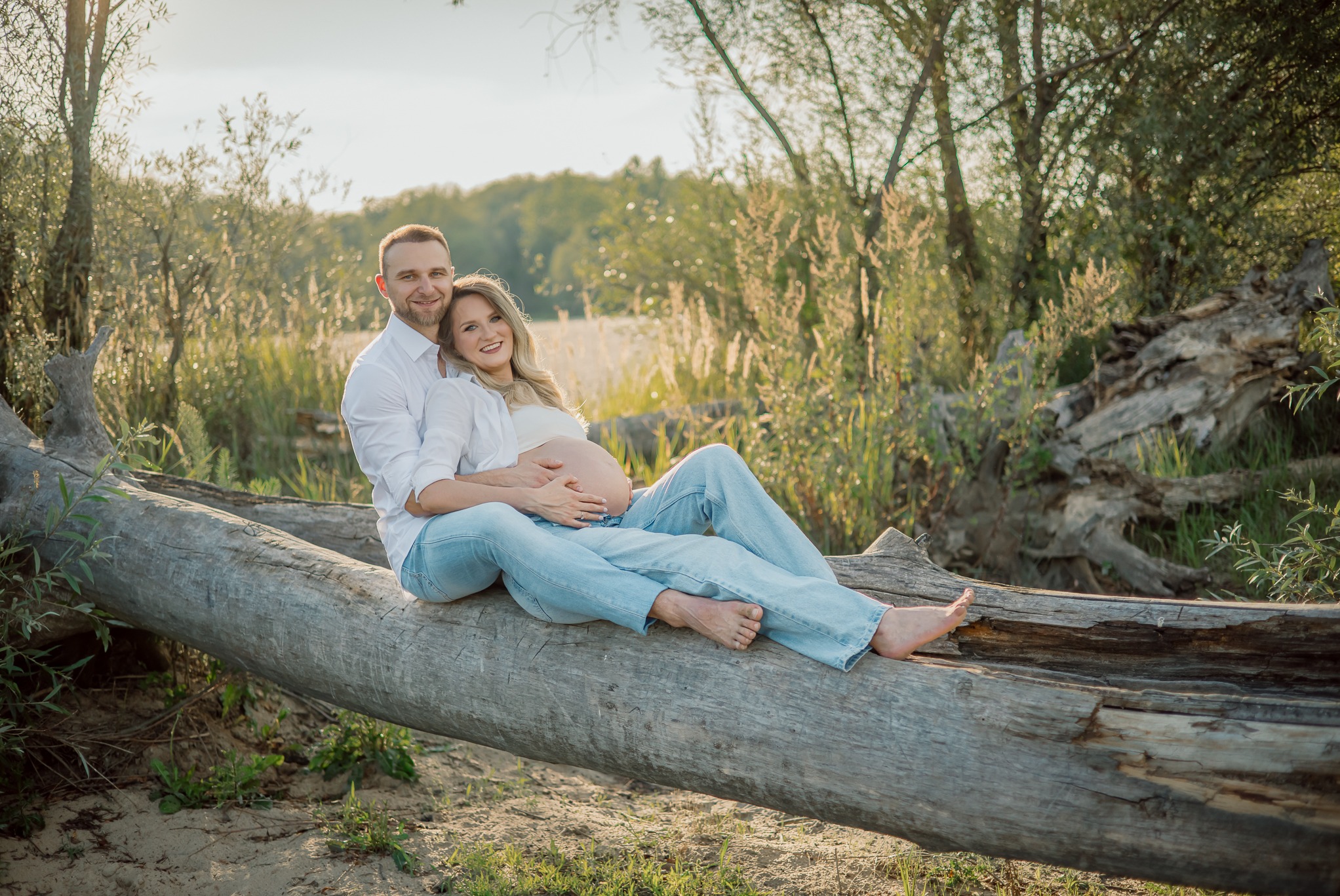 Para w jasnych ubraniach siedzi na powalonym drzewie w plenerze. Kobieta w ciąży, mężczyzna obejmuje ją. Romantyczna sesja w naturalnym otoczeniu, w ciepłym świetle.