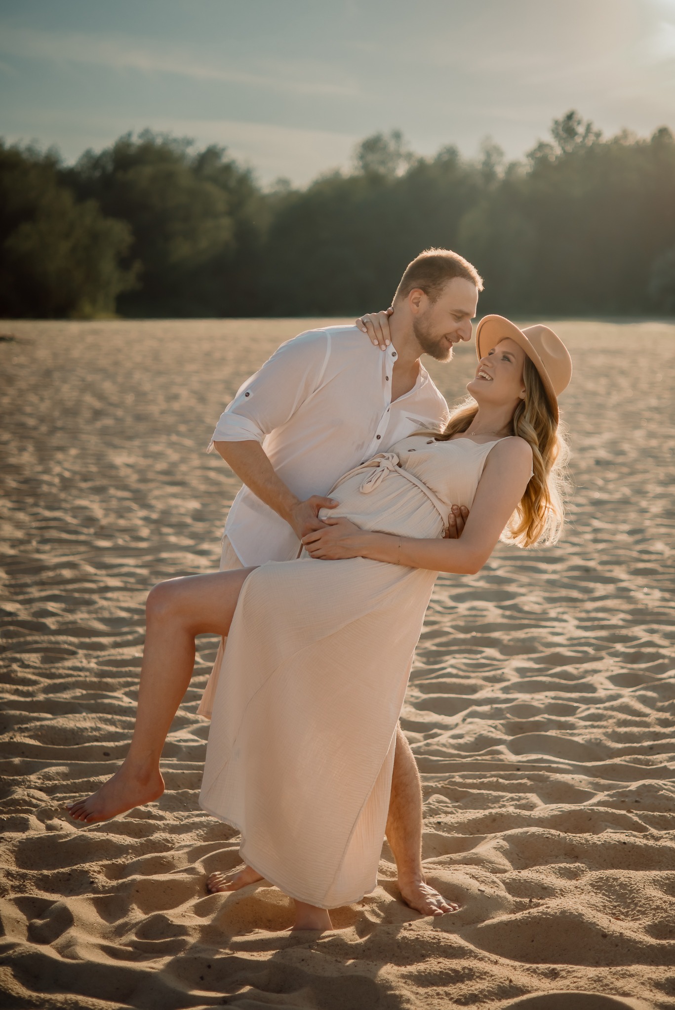 Para na plaży w ciepłym świetle: mężczyzna trzyma uśmiechniętą, ciężarną kobietę w zwiewnej sukience i kapeluszu. Romantyczna sesja ciążowa na tle piaszczystej plaży i drzew.