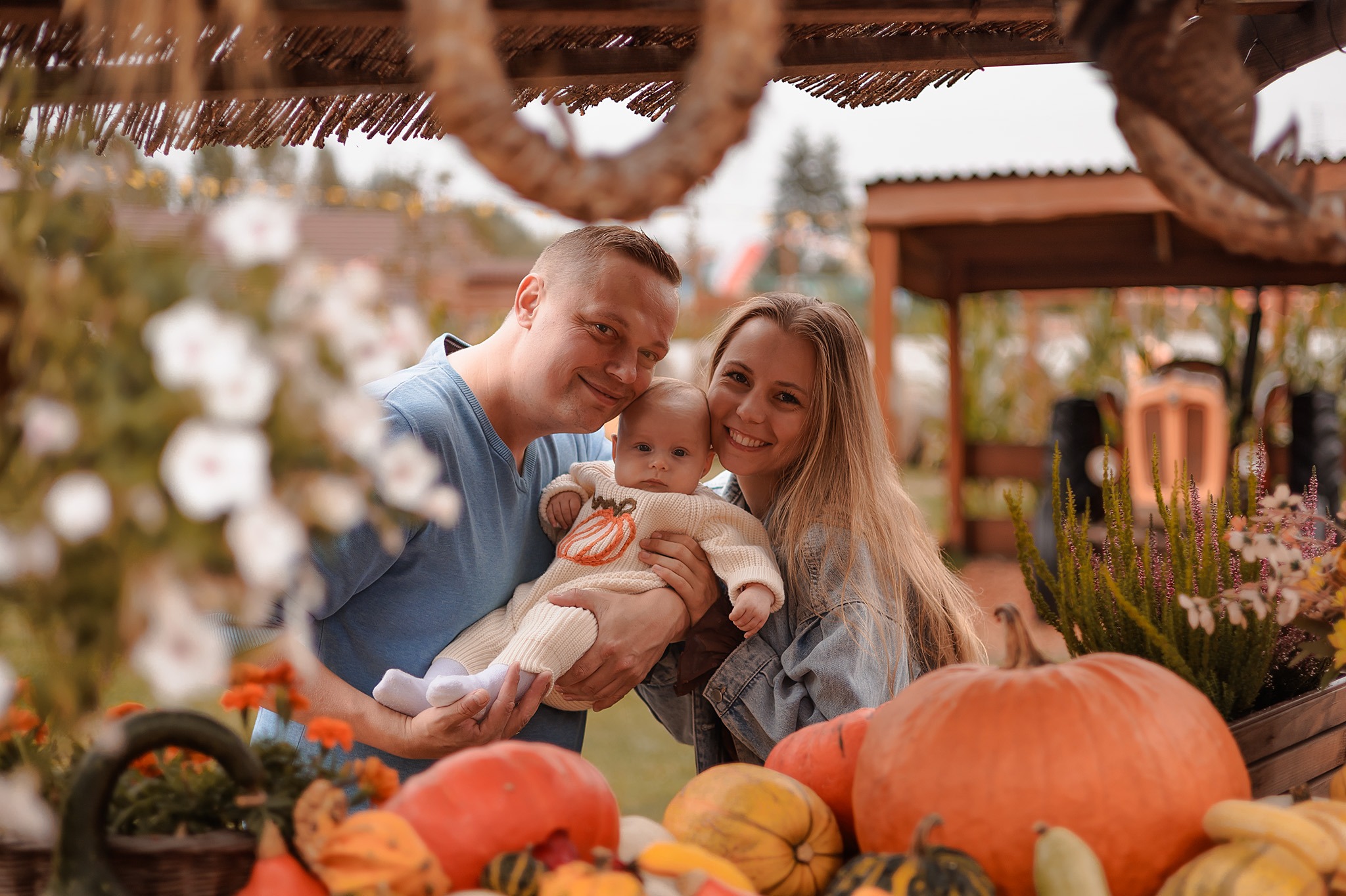 Jesienna sesja rodzinna: uśmiechnięta rodzina z niemowlęciem w dyniowym sweterku pozuje wśród dyń i kwiatów, na tle rustykalnej scenerii.