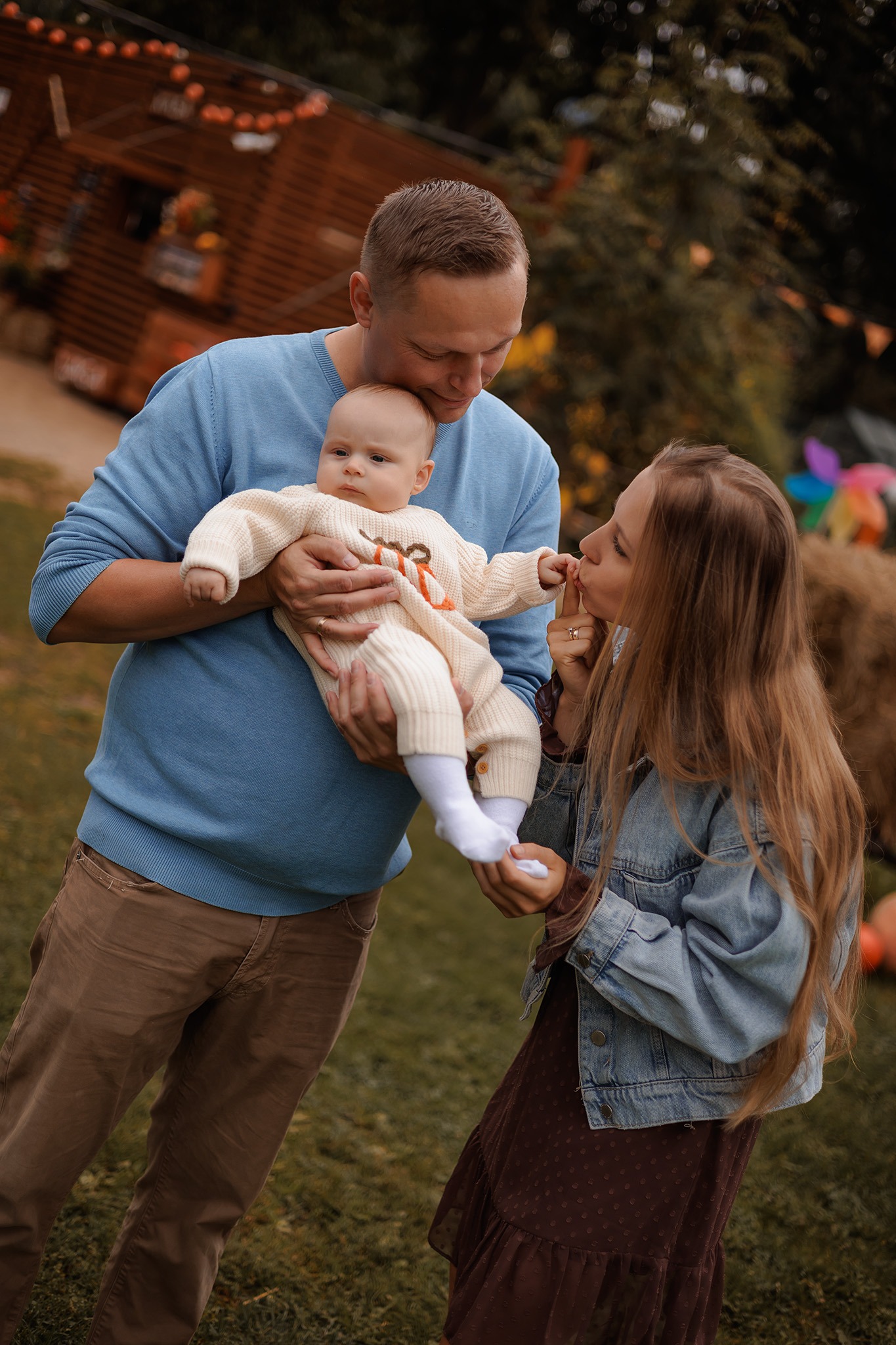 Jesienna sesja rodzinna: ojciec trzyma niemowlę w kremowym kombinezonie, matka całuje rączkę dziecka, w tle dekoracje z dyni i drewniany domek.