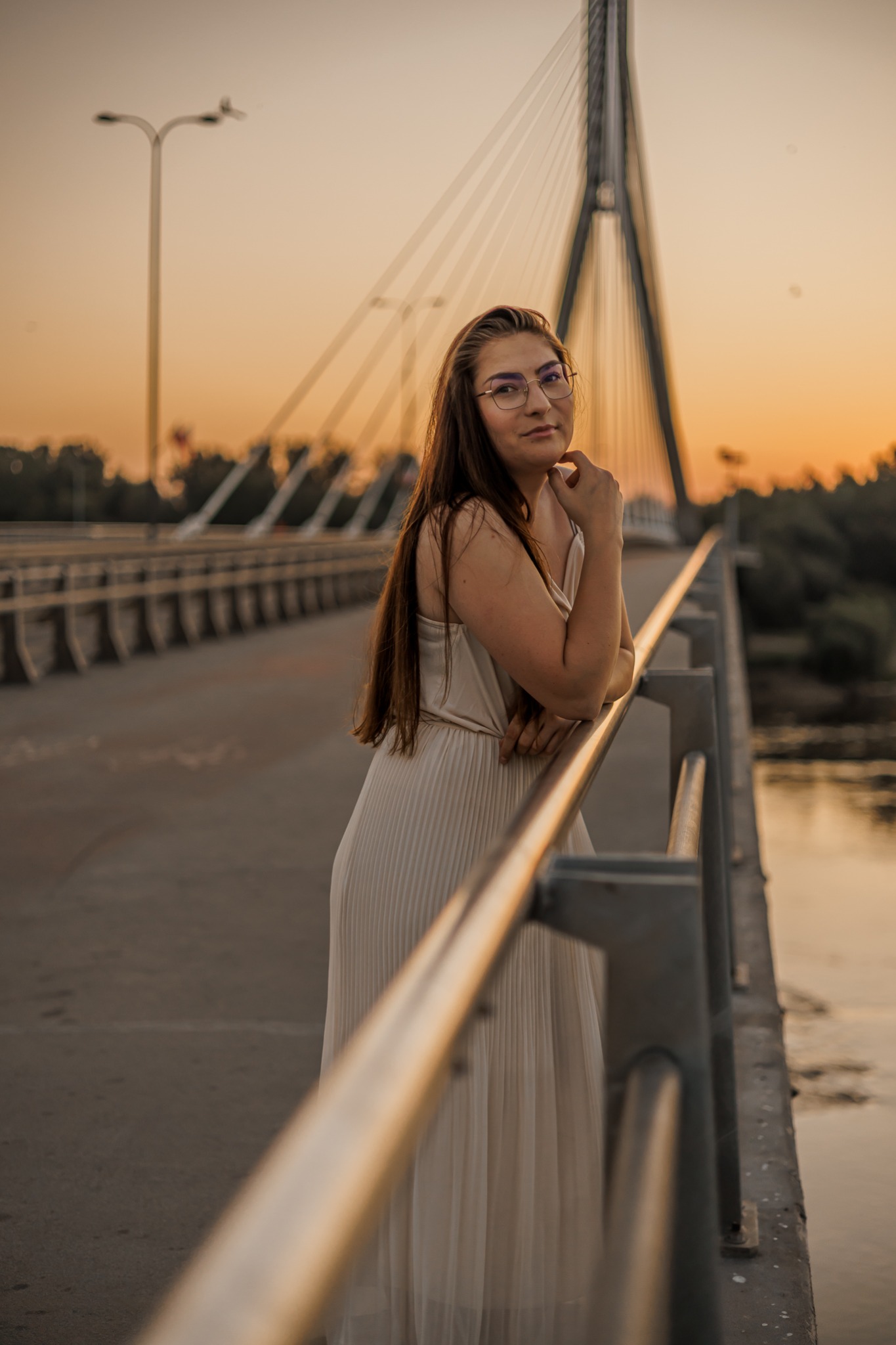 Portret kobiety w okularach, opierającej się o barierkę mostu w promieniach zachodzącego słońca. W tle widoczna konstrukcja mostu i rzeka.