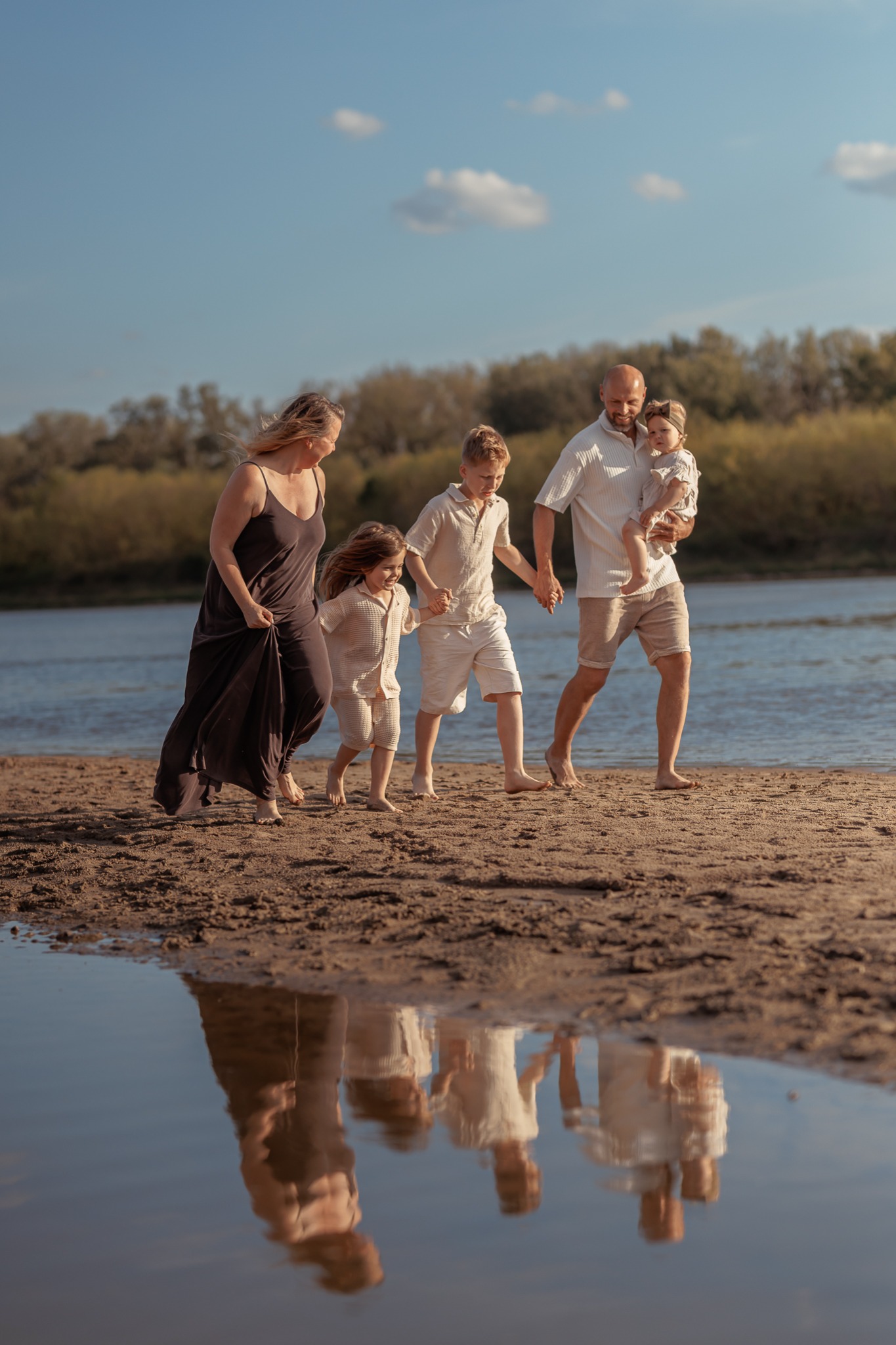 Rodzina z trójką dzieci spaceruje brzegiem rzeki, trzymając się za ręce. Odbicie w wodzie dodaje dynamiki. Sesja rodzinna w naturalnym otoczeniu.