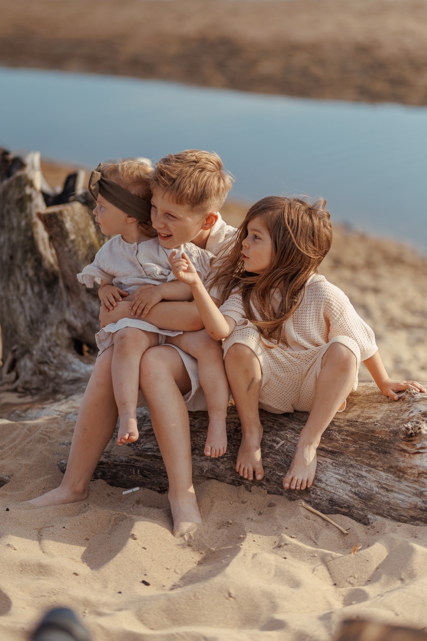 Trójka uroczych dzieci w beżowych ubraniach siedzi na pniu drzewa na piaszczystej plaży z widokiem na wodę w tle. Zdjęcie w ciepłych, naturalnych barwach.