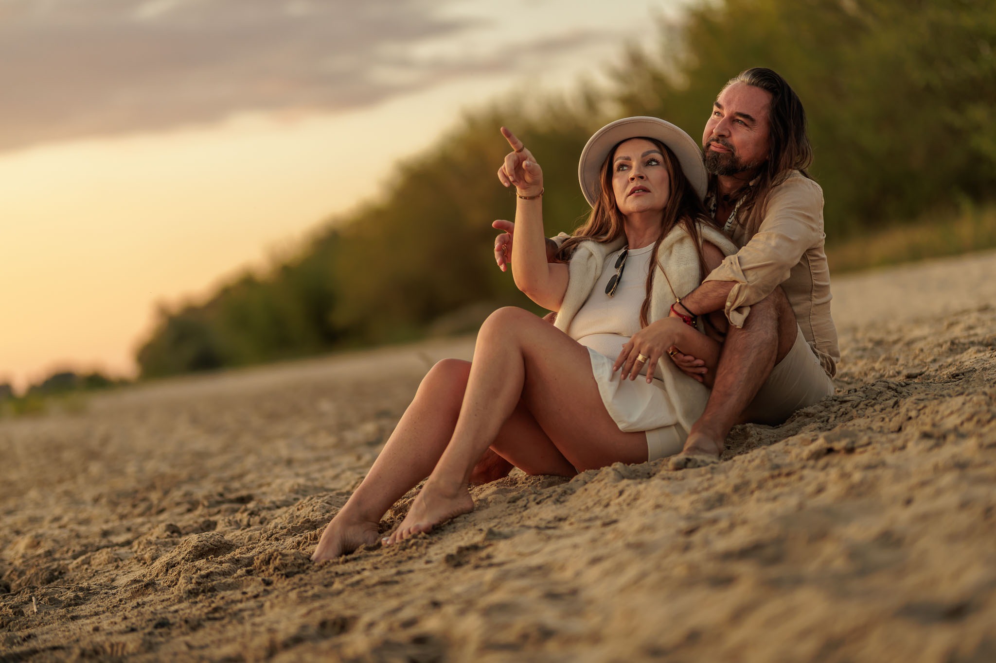 Para siedzi na piasku o zachodzie słońca. Kobieta w kapeluszu wskazuje palcem w górę, mężczyzna obejmuje ją ramionami, oboje patrzą w tym samym kierunku.