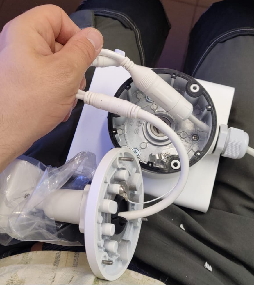 Montaż kamery monitoringu: dłoń łączy przewody do podstawy kamery. Widoczna obudowa, kable i elementy montażowe na tle ubrania.