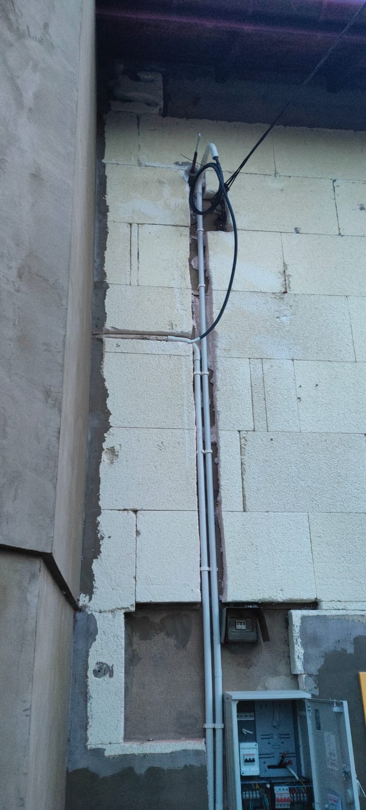 Instalacja elektryczna zewnętrzna: przewody w peszlu, licznik i skrzynka rozdzielcza na elewacji budynku z bloków. Widoczne kable i rury pionowe.
