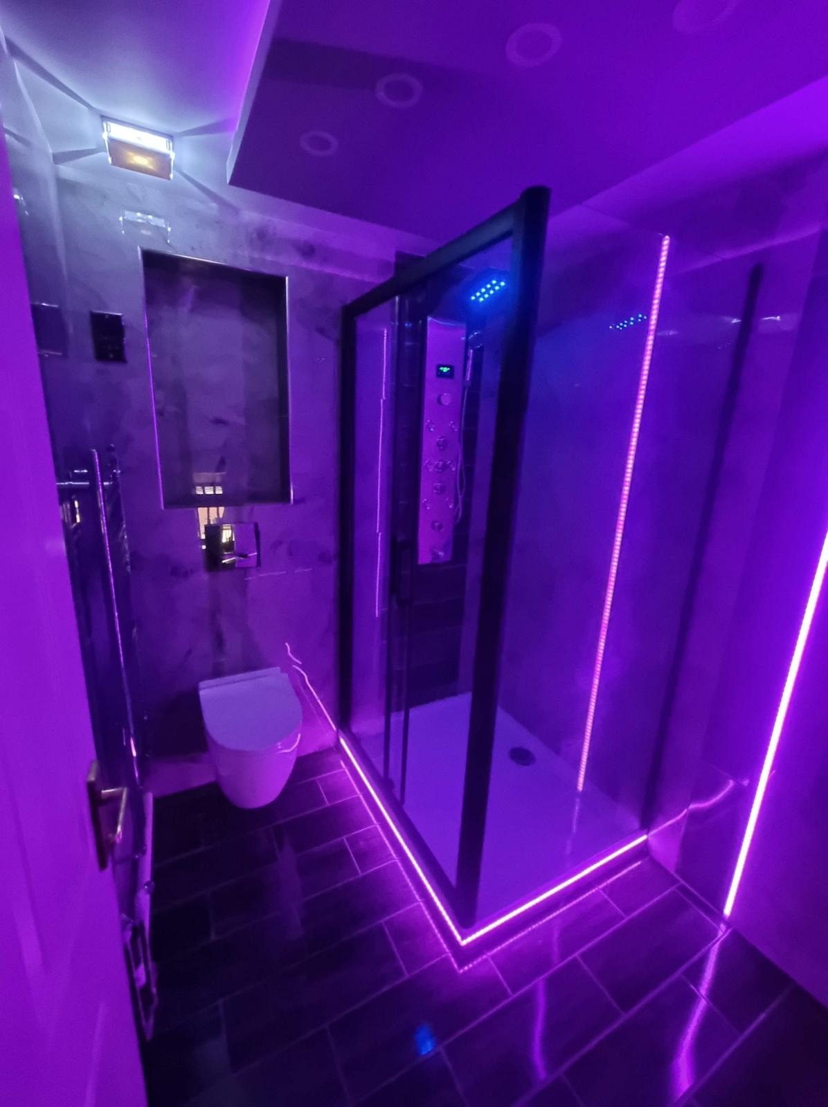 Nowoczesna łazienka z czarną kabiną prysznicową, podświetleniem LED w kolorze fioletowym i szarą glazurą. Uwagę przykuwa minimalistyczna toaleta i lustro.