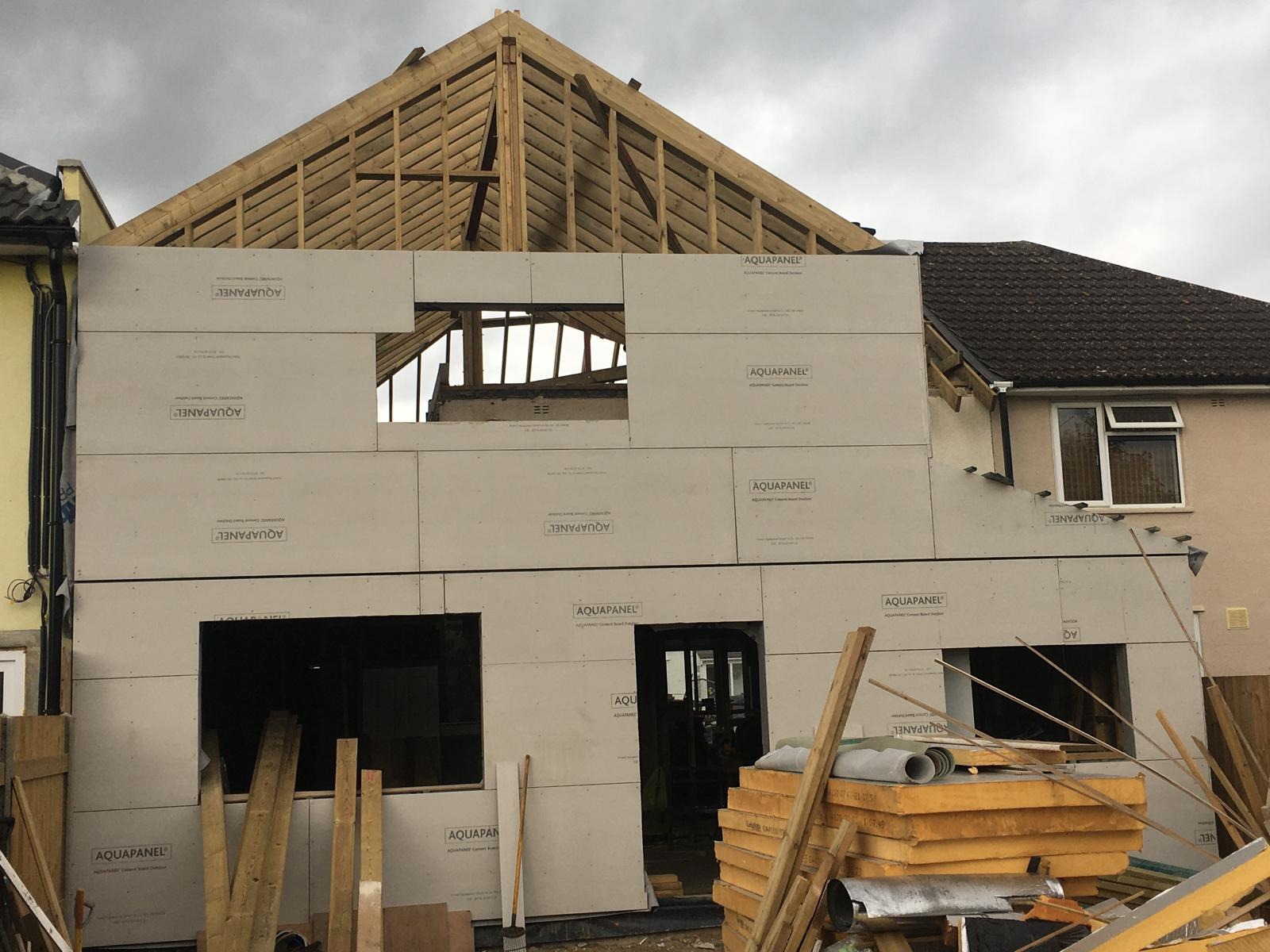 Montaż płyt Aquapanel na elewacji domu jednorodzinnego. Widoczne drewniane elementy konstrukcji dachu i okna. Częściowo wykonane ocieplenie budynku.