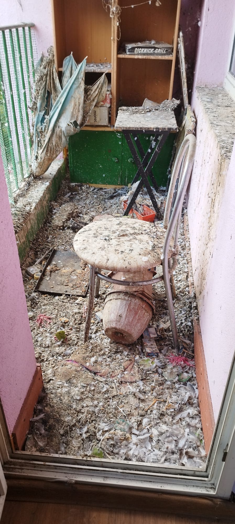 Zaniedbany balkon z meblami pokrytymi ptasimi odchodami i piórami. Widoczny regał, stolik, krzesło i doniczka. Silne zabrudzenie, brak dbałości o przestrzeń.