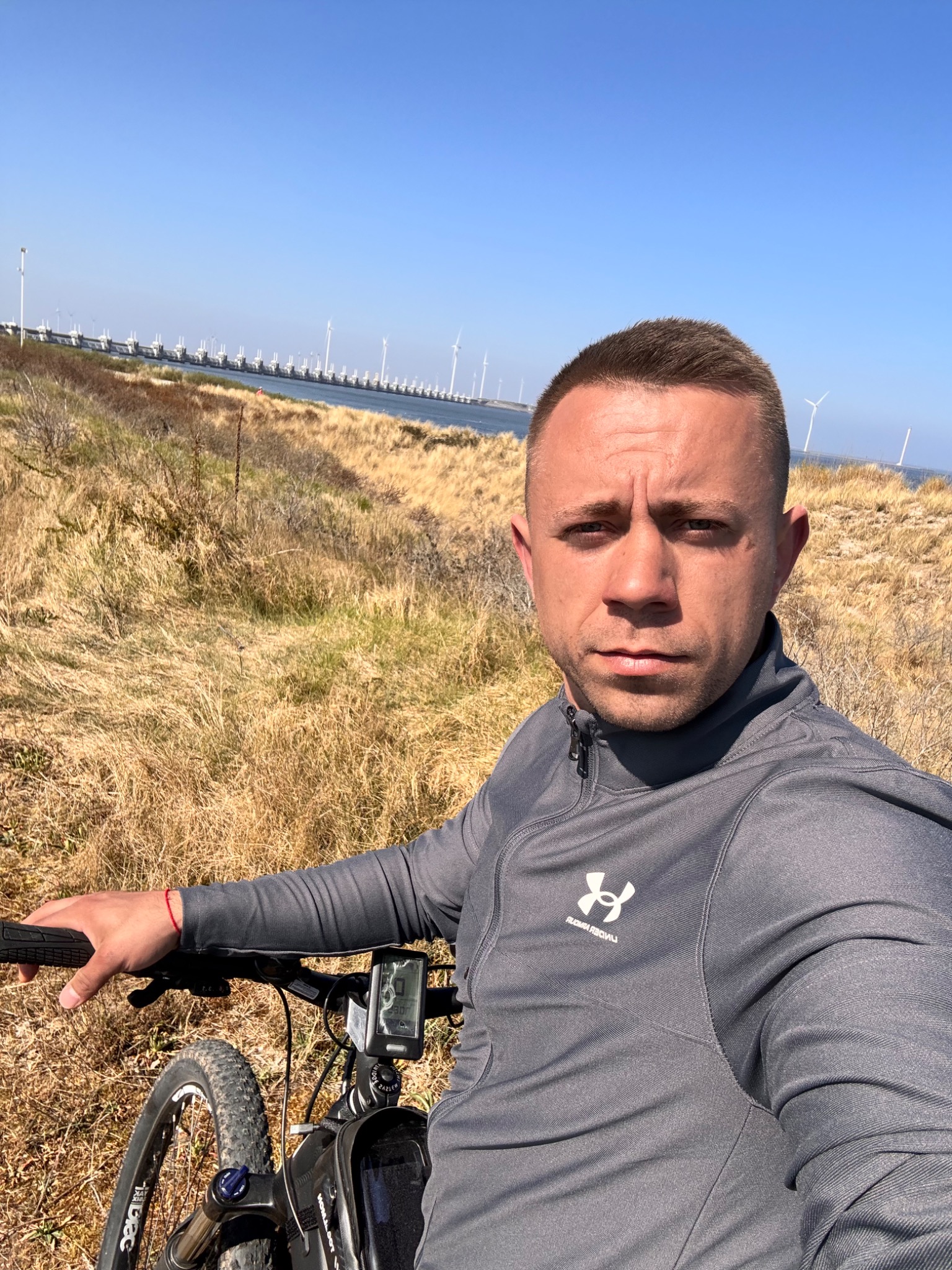 Mężczyzna w szarej bluzie na rowerze, w tle farma wiatrowa i błękitne niebo. Autoportret w plenerze, bliska odległość od obiektywu.