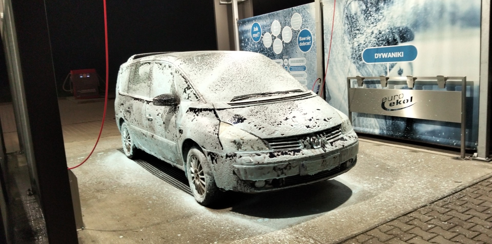 Samochód typu minivan pokryty pianą w myjni bezdotykowej, widoczny automat i stanowisko na dywaniki. Nocne oświetlenie.