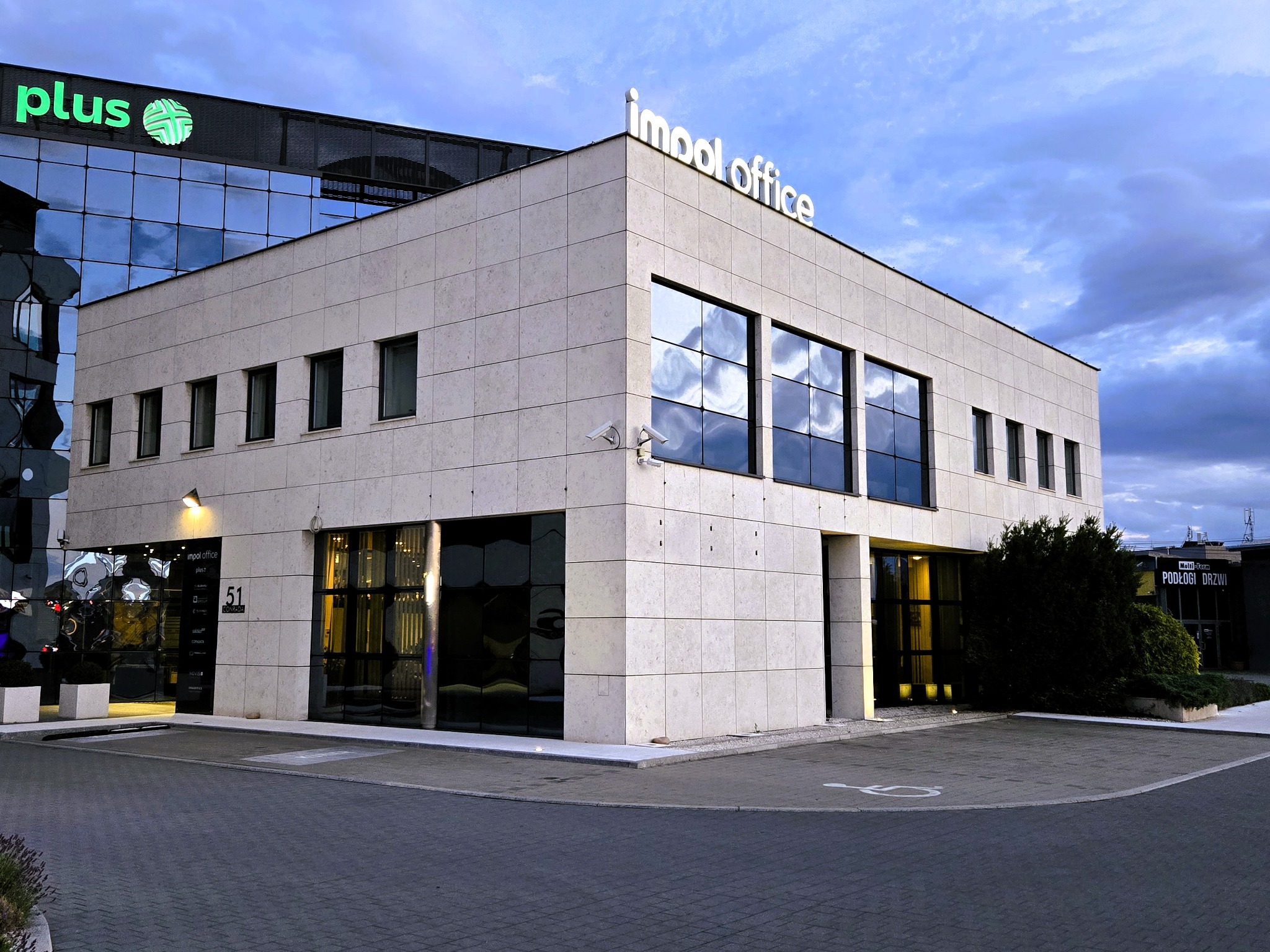 Nowoczesny biurowiec z logo 'impol office' i 'plus' na fasadzie, widok z poziomu ulicy, kostka brukowa, znak dla niepełnosprawnych na parkingu.