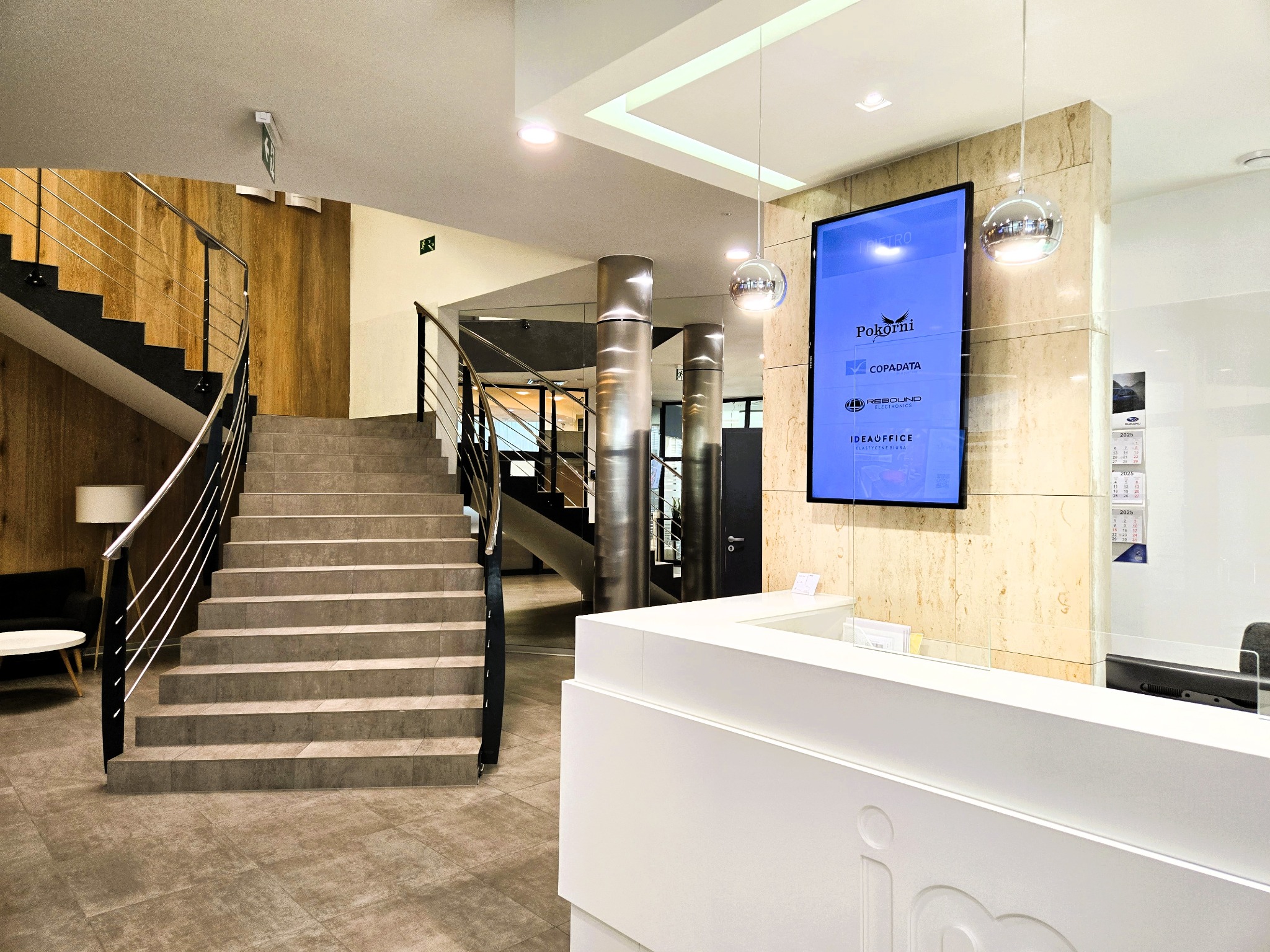 Recepcja w nowoczesnym biurze z marmurową ścianą, schodami i wiszącymi lampami. Na ścianie logo firm Pokorni, Copadata, Rebound i IdeaOffice.