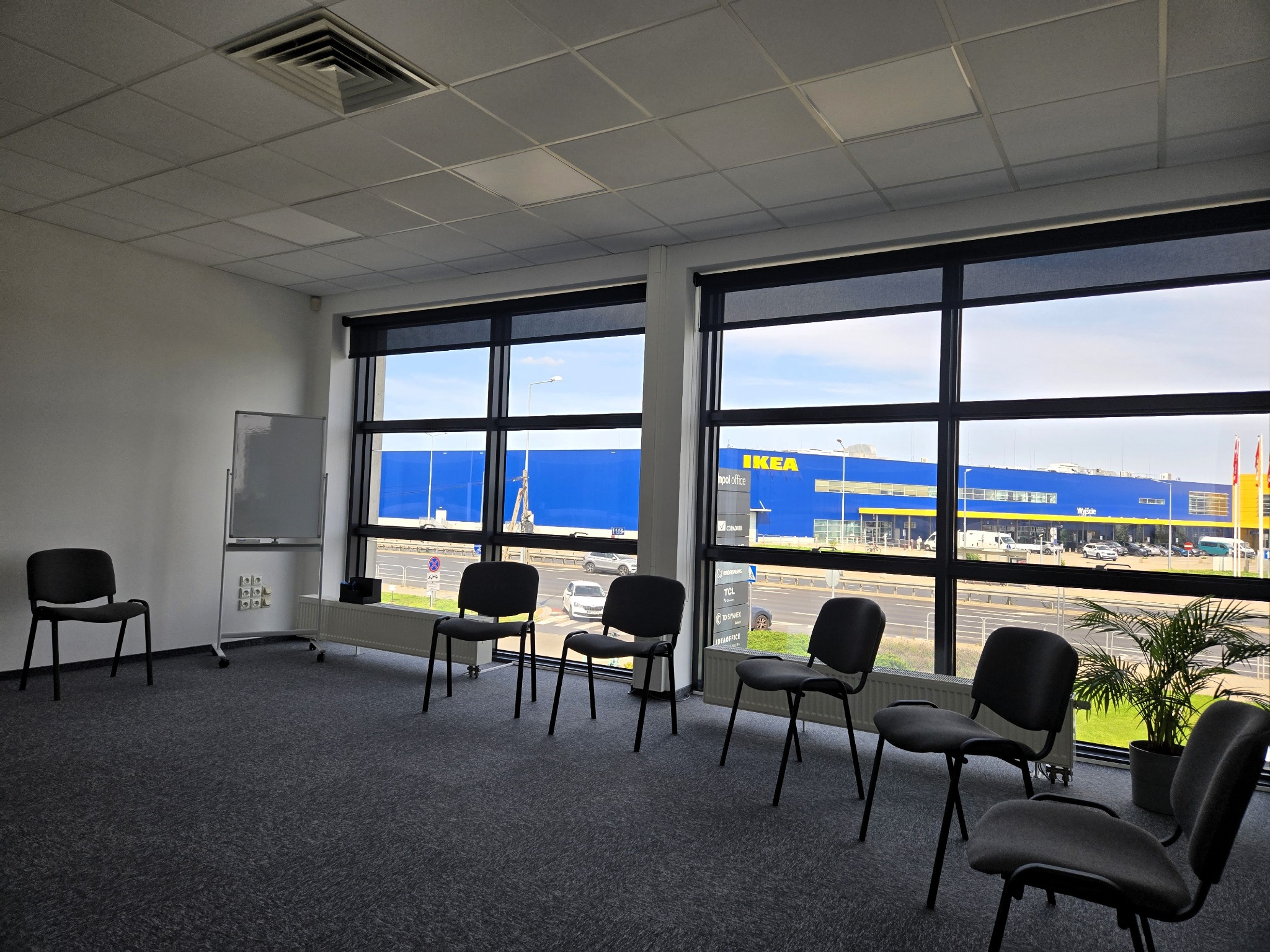 Sala konferencyjna z krzesłami i tablicą, widok na budynek IKEA przez okno. Szara wykładzina, białe ściany, rolety częściowo zasłonięte. Dzień.