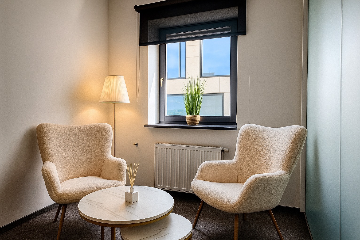 Jasny pokój terapii z dwoma fotelami typu boucle, stolikiem kawowym z dyfuzorem zapachowym i widokiem na budynek przez okno z roletą.