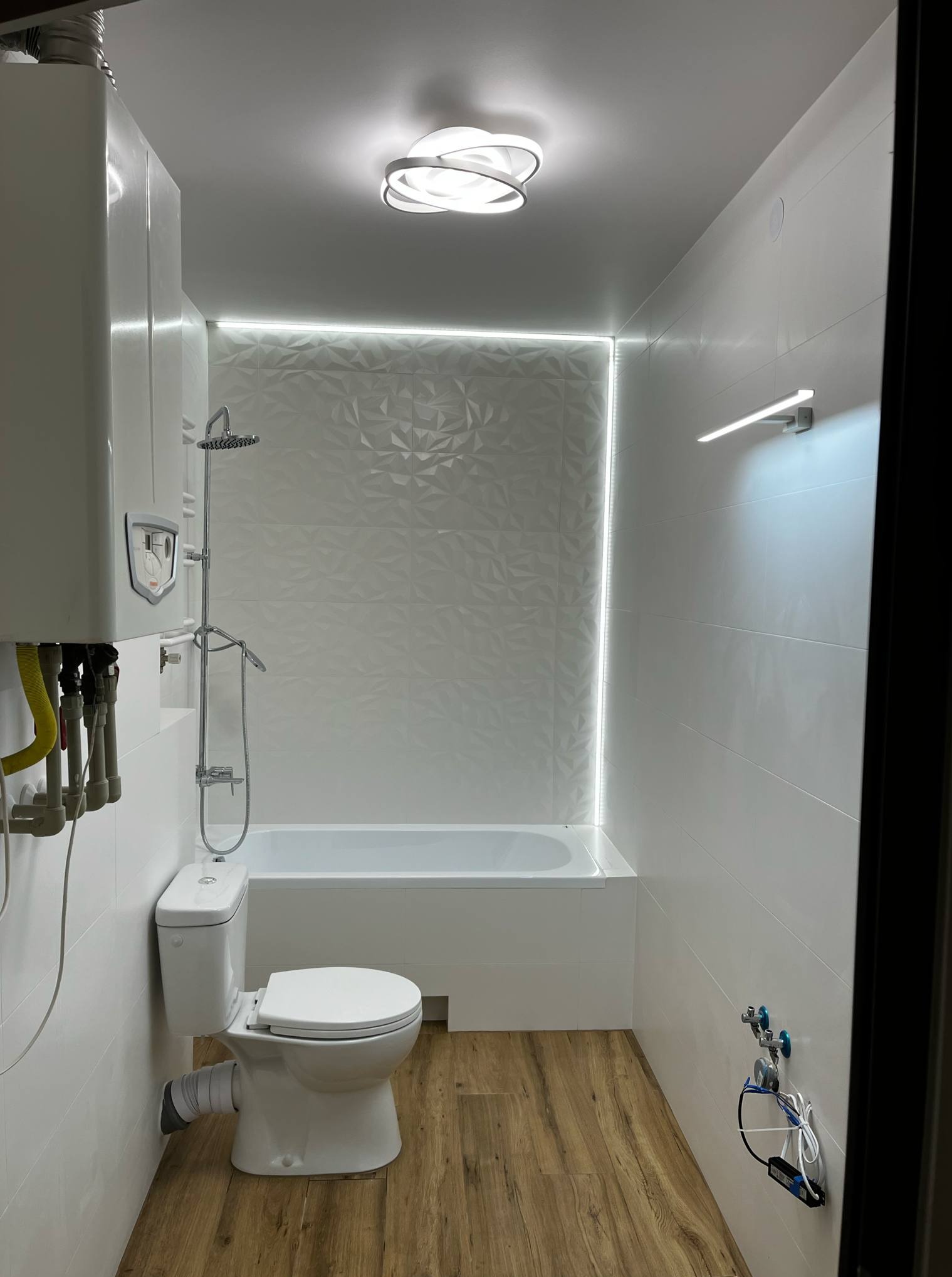 Nowoczesna łazienka po remoncie: białe płytki 3D, wanna z prysznicem, podświetlenie LED, drewnopodobna podłoga i designerska lampa sufitowa.