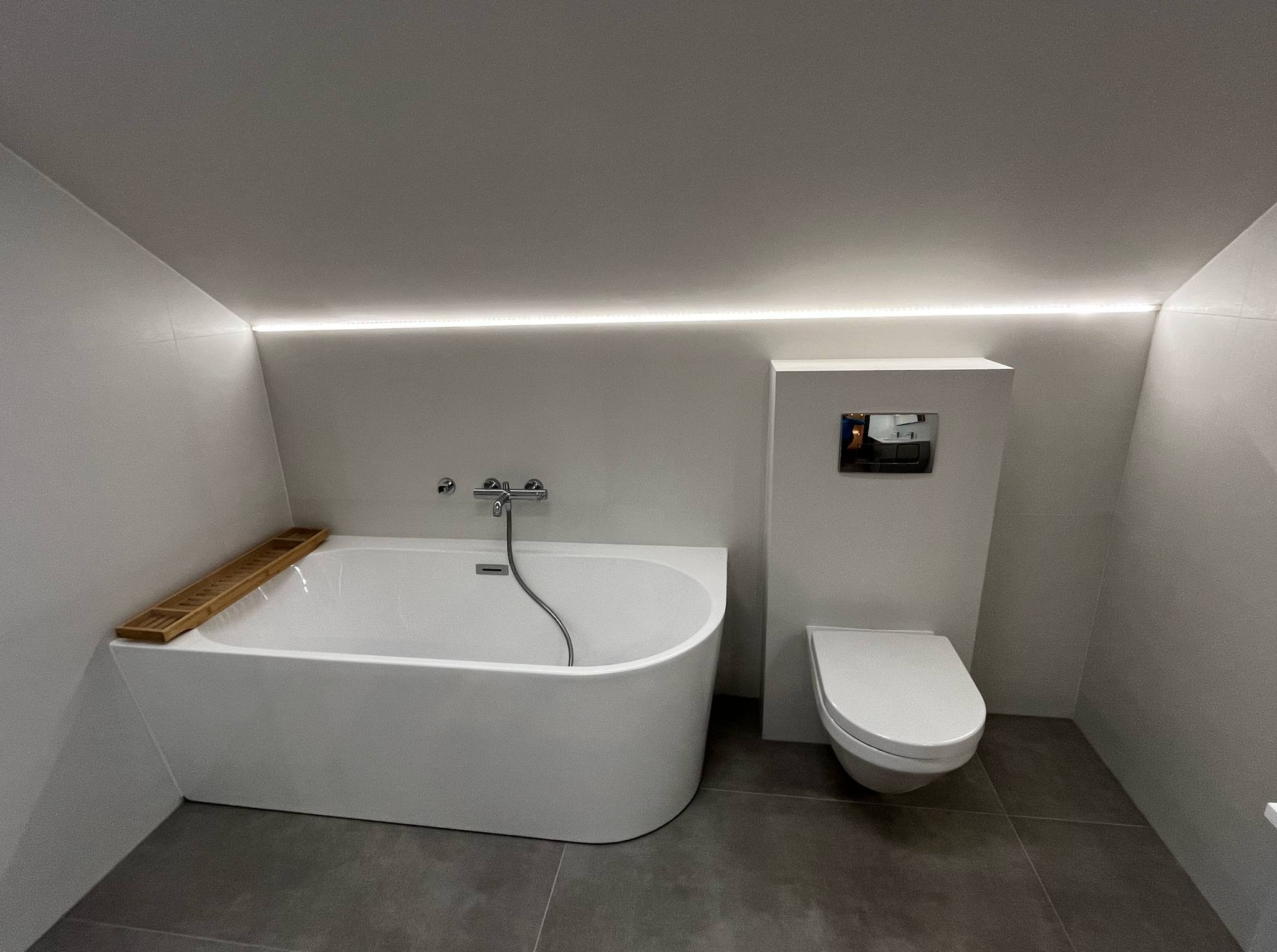 Nowoczesna łazienka na poddaszu z wanną, toaletą podtynkową i ledowym oświetleniem sufitowym. Minimalistyczny design, szare płytki podłogowe.