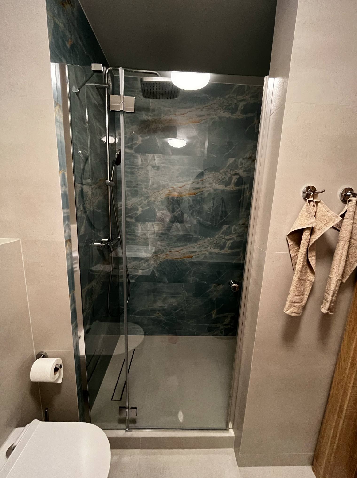 Nowoczesna kabina prysznicowa z deszczownicą i marmurowymi płytkami w odcieniach zieleni i złota. Na ścianie wieszaki z ręcznikami w neutralnych barwach.