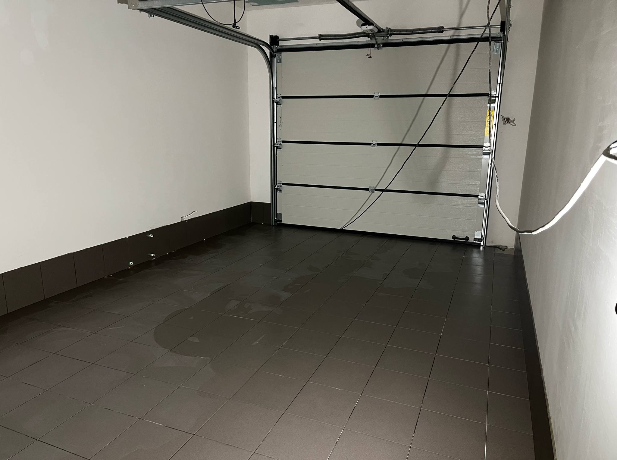 Wykończone wnętrze garażu z ciemnymi, błyszczącymi płytkami podłogowymi i segmentową bramą garażową. Widoczne listwy przypodłogowe i gładkie, białe ściany.