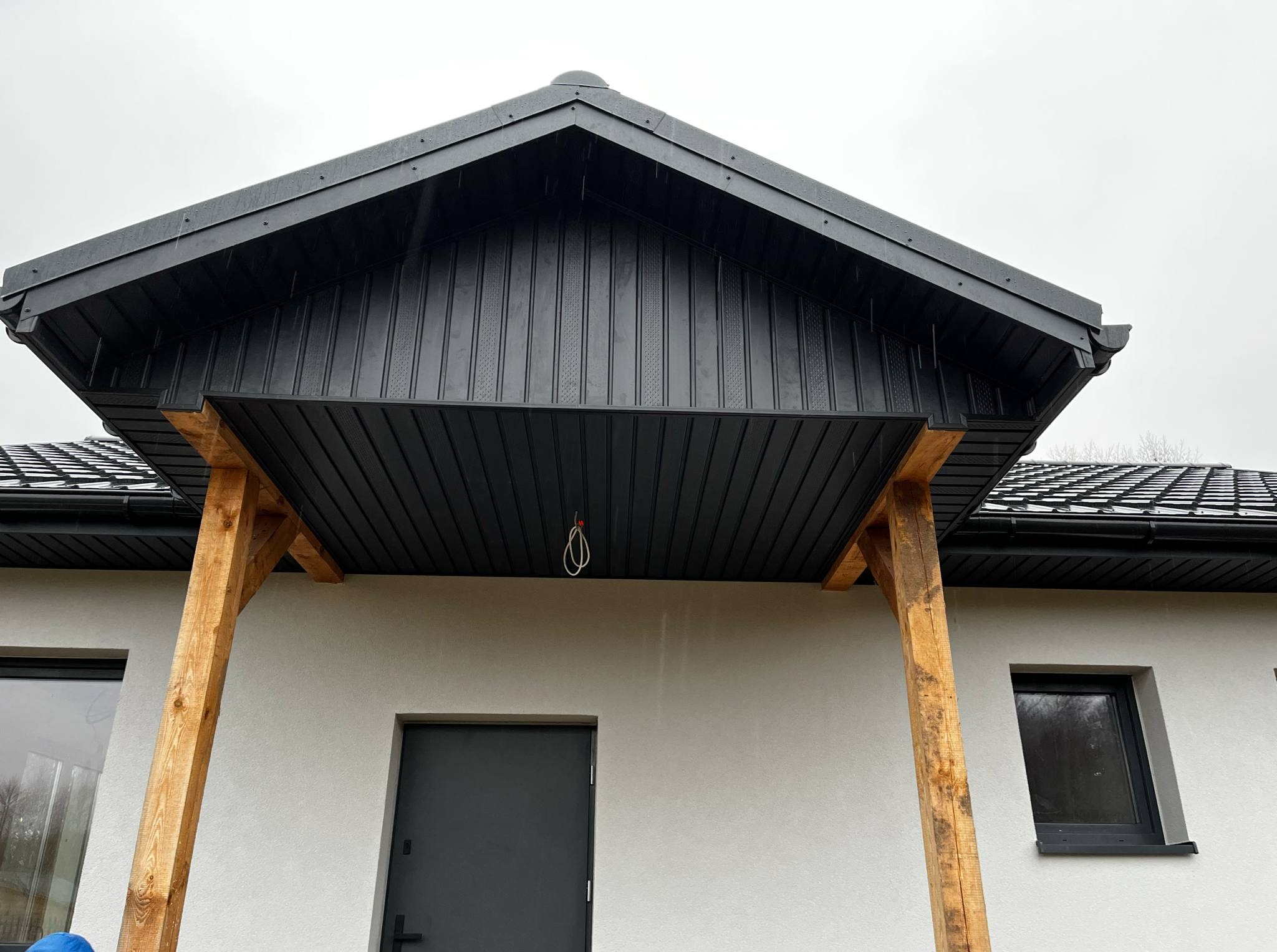 Nowoczesny, minimalistyczny dom z czarnym zadaszeniem nad wejściem, wspartym na drewnianych belkach. Szara elewacja, czarne okna i drzwi.