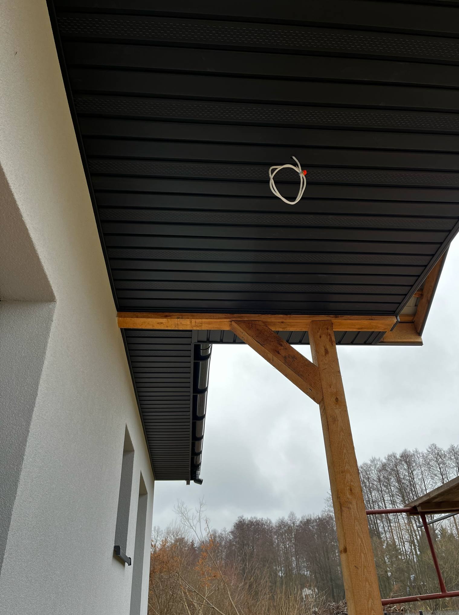 Czarne panele podsufitowe zamontowane na zewnątrz budynku, widoczne drewniane elementy konstrukcyjne i wystający przewód elektryczny.
